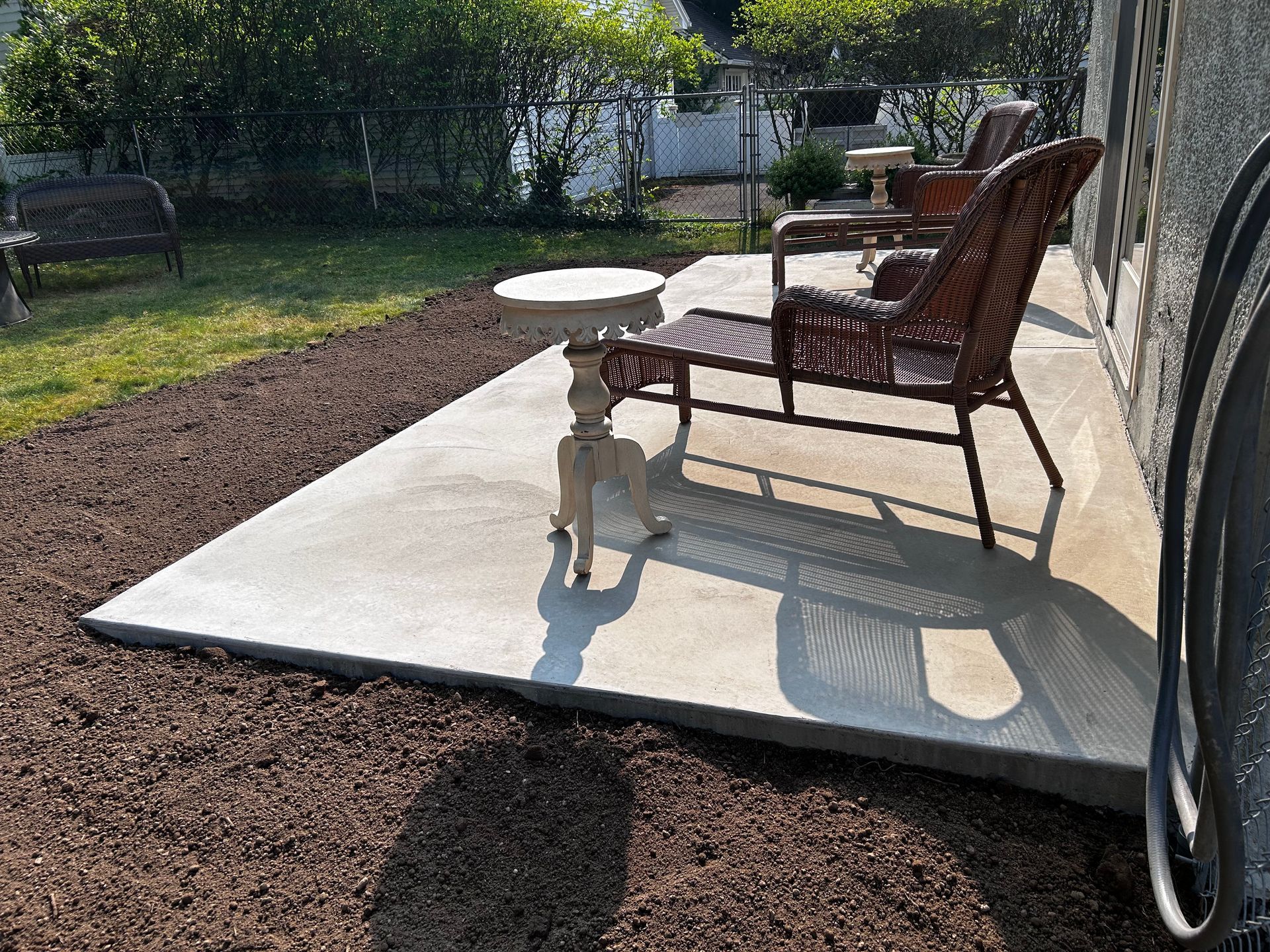 A concrete patio with two wicker chairs and a small white side table, surrounded by dark mulch in a backyard setting.
