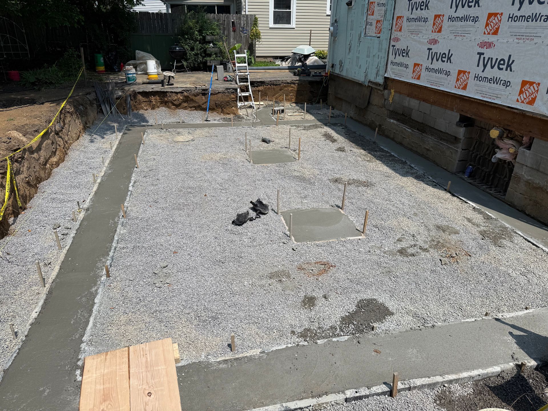 A gravel-filled construction site with concrete foundation footings and trenches for a new building extension.