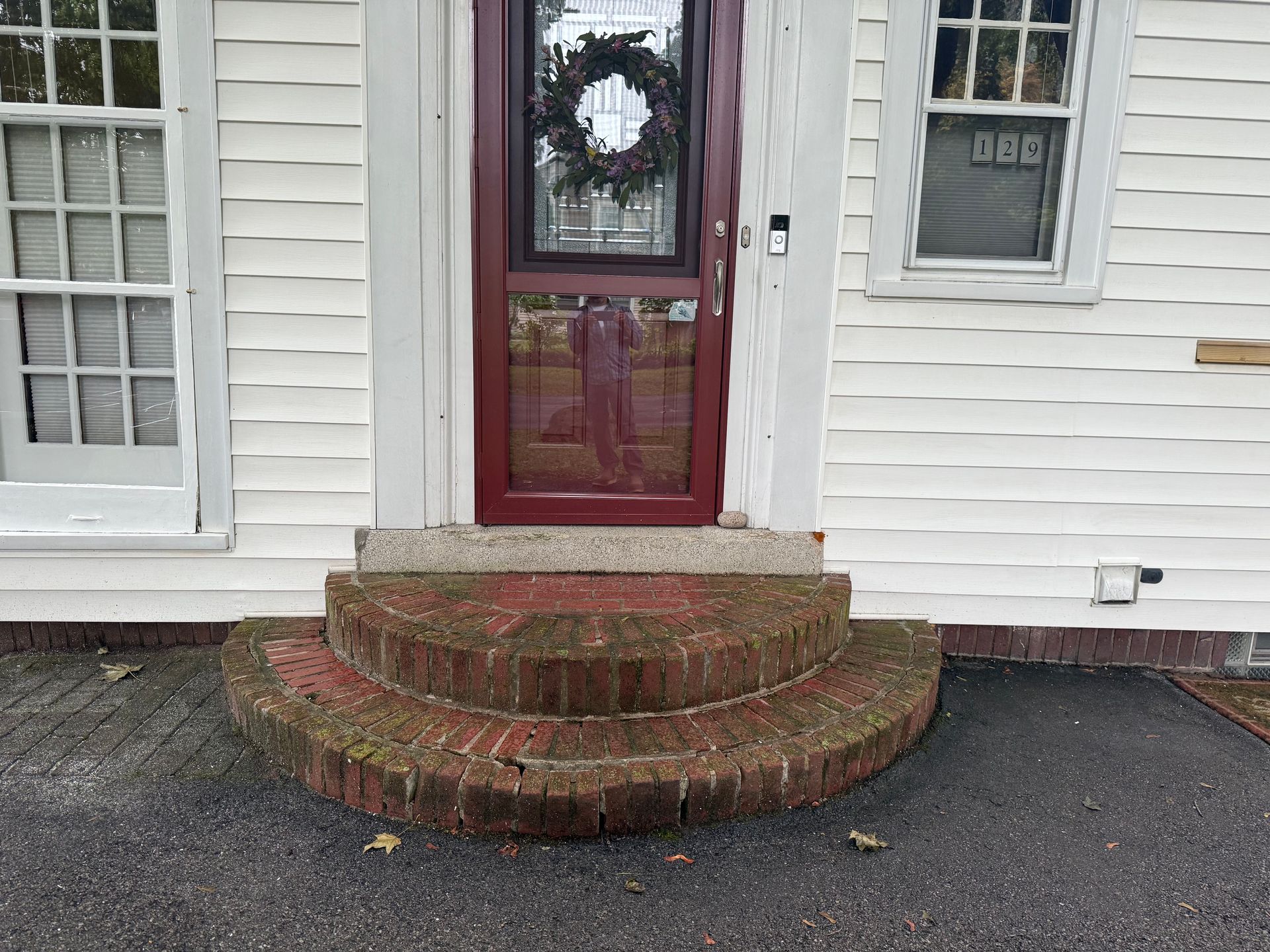 A red-trimmed front door with a wreath, set above two semicircular brick steps on a white-sided house.