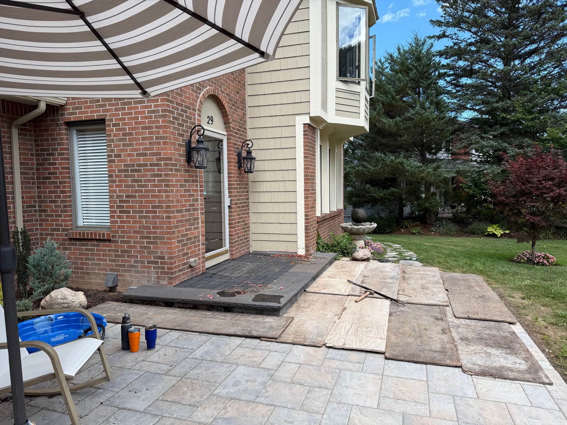 A patio under construction with brick pavers, a row of new concrete slabs, and a house exterior with brick and siding.