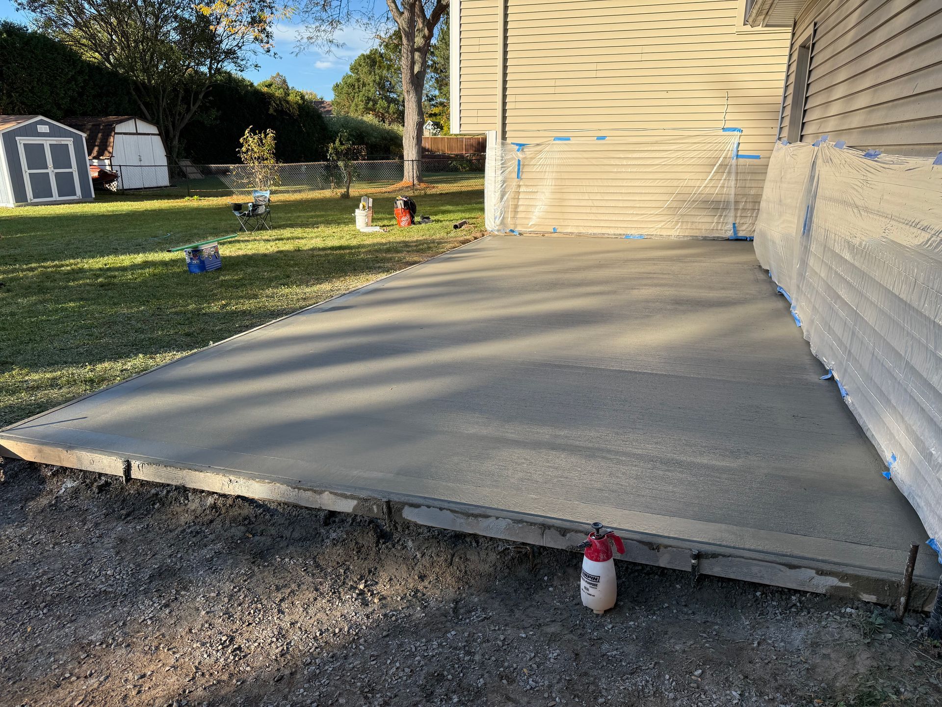 A freshly poured concrete slab adjacent to a house, with a lawn, storage shed, and spray bottle in the foreground.