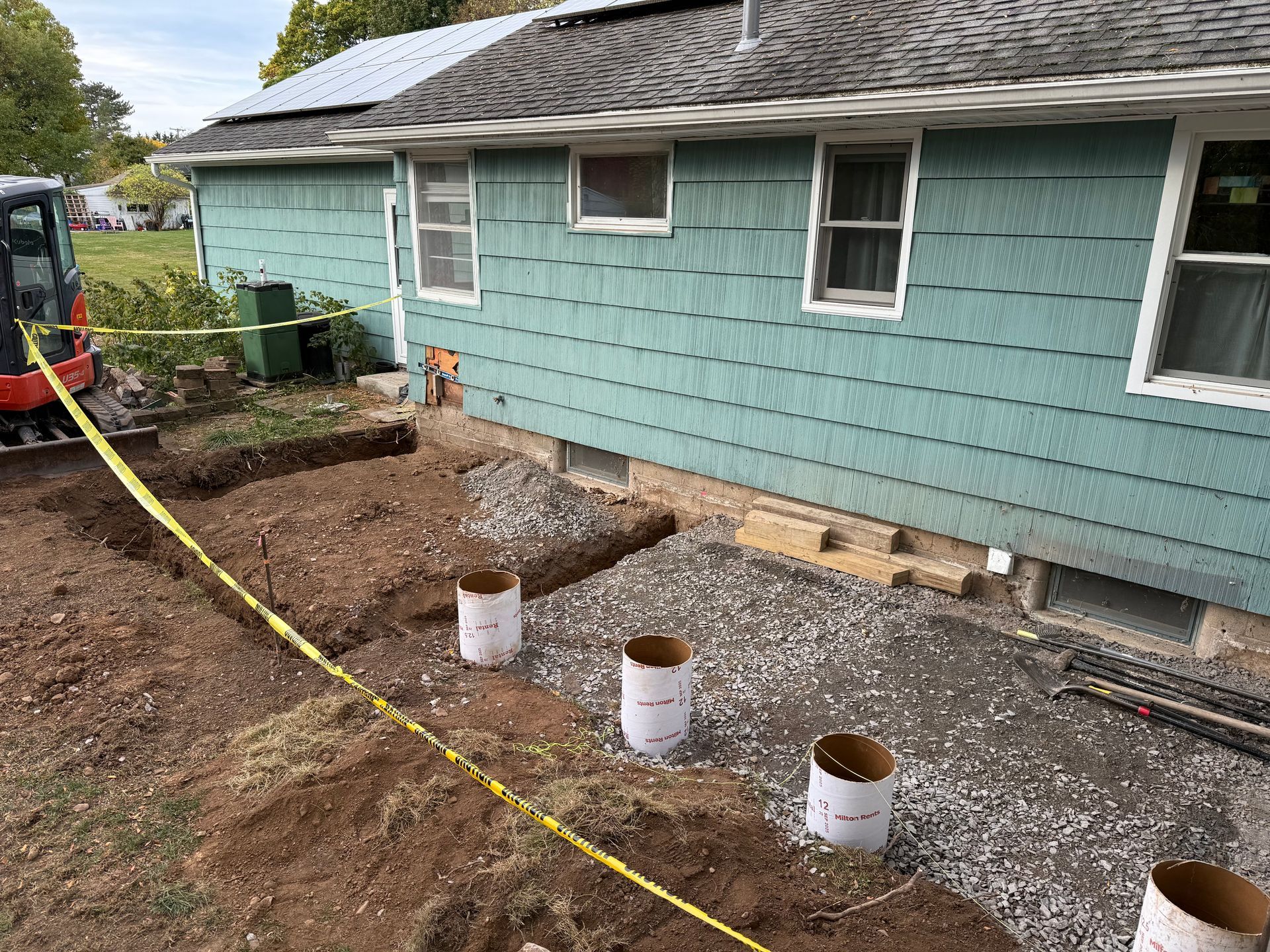 A construction site at the back of a blue house, showing a gravel base with several circular concrete forms for a deck.