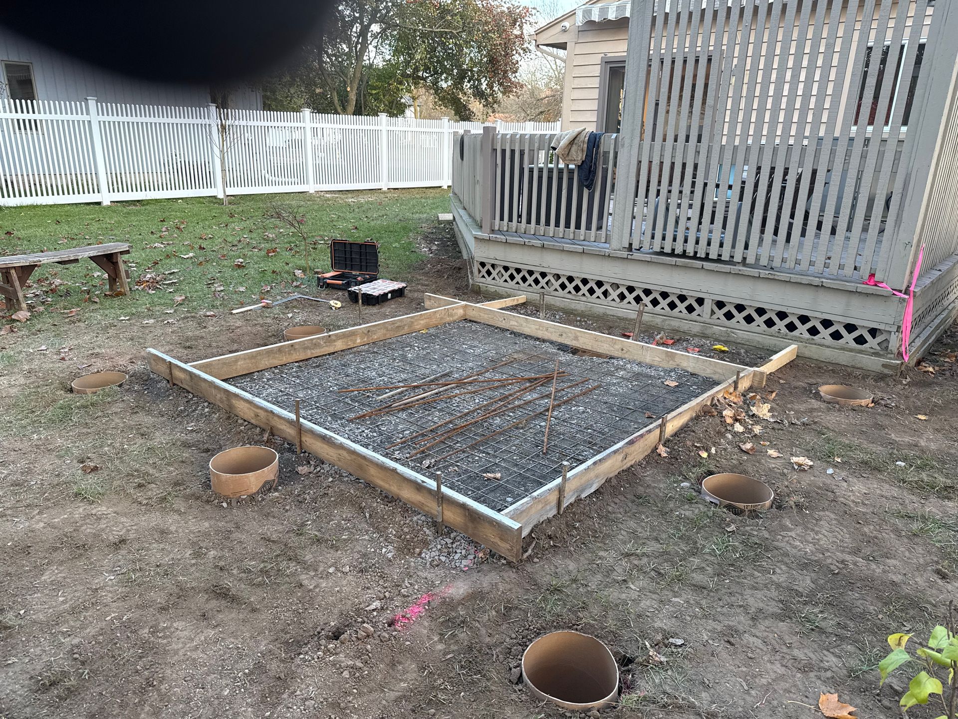 A square wooden concrete form filled with gravel sits in a yard next to a deck, with empty concrete pier holes in the ground.