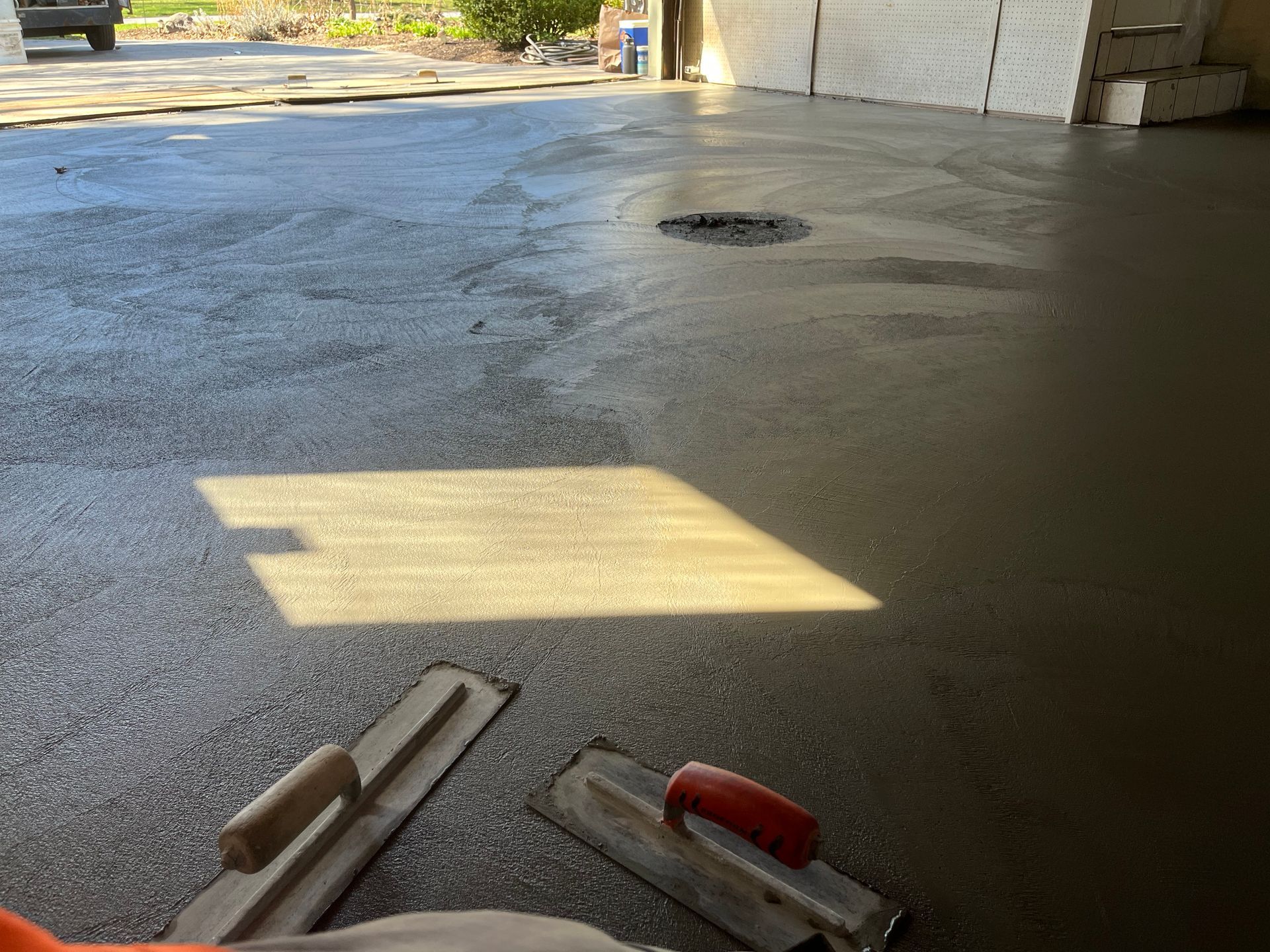 Two hand trowels lie on a freshly poured, wet concrete floor inside a garage.