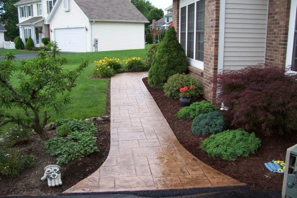 A stamped concrete walkway leads to the front entrance of a brick house, flanked by green landscaping and mulch beds.
