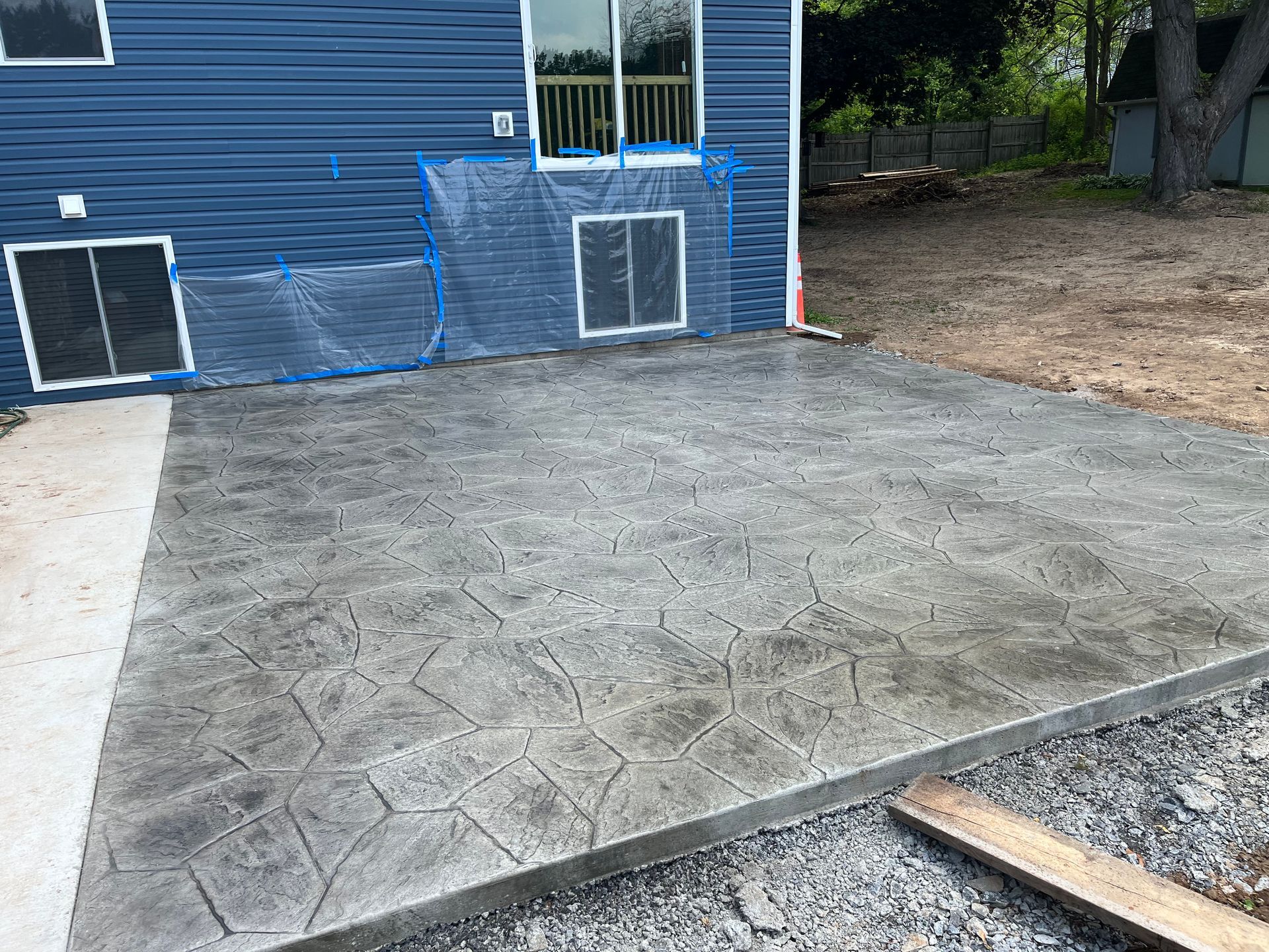 A newly installed grey stamped concrete patio outside a dark blue house with two ground-level windows.