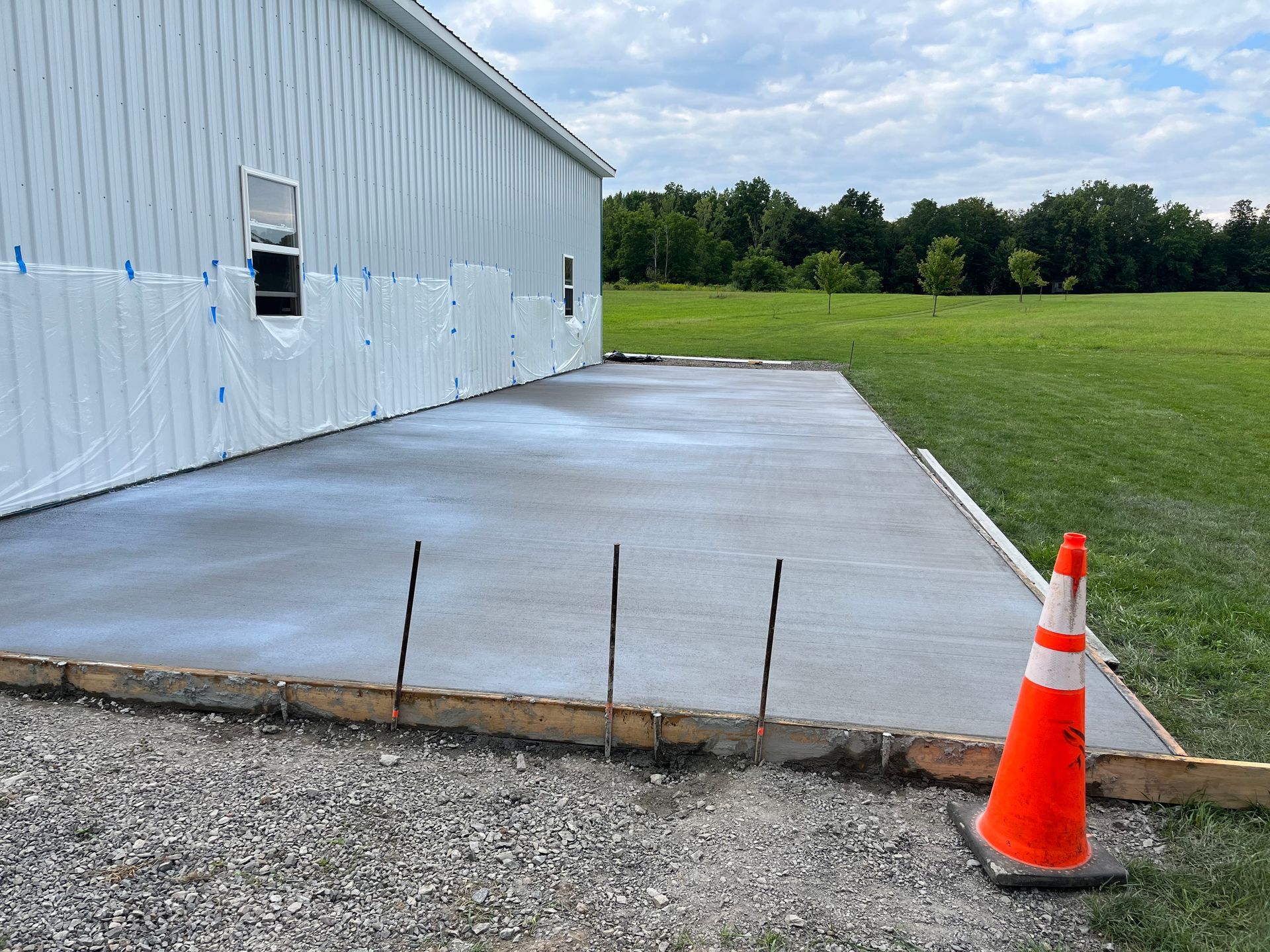Freshly poured concrete slab extending from the side of a metal-sided building, with a traffic cone on the gravel.
