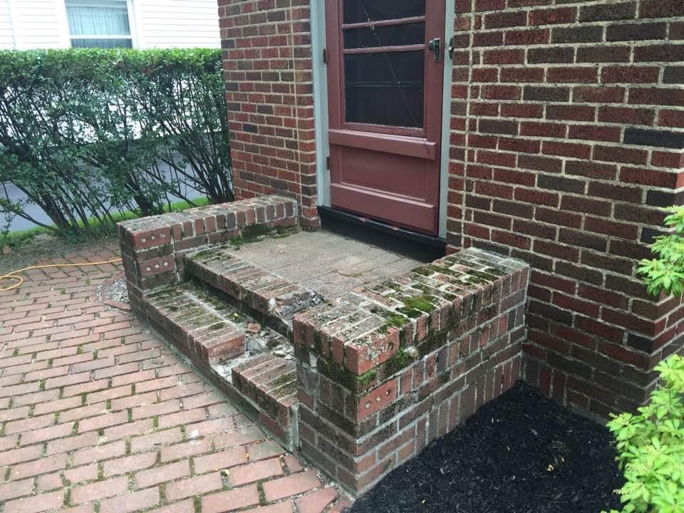 A brick entryway with two steps leading to a door, showing signs of moss and deterioration on a brick patio.