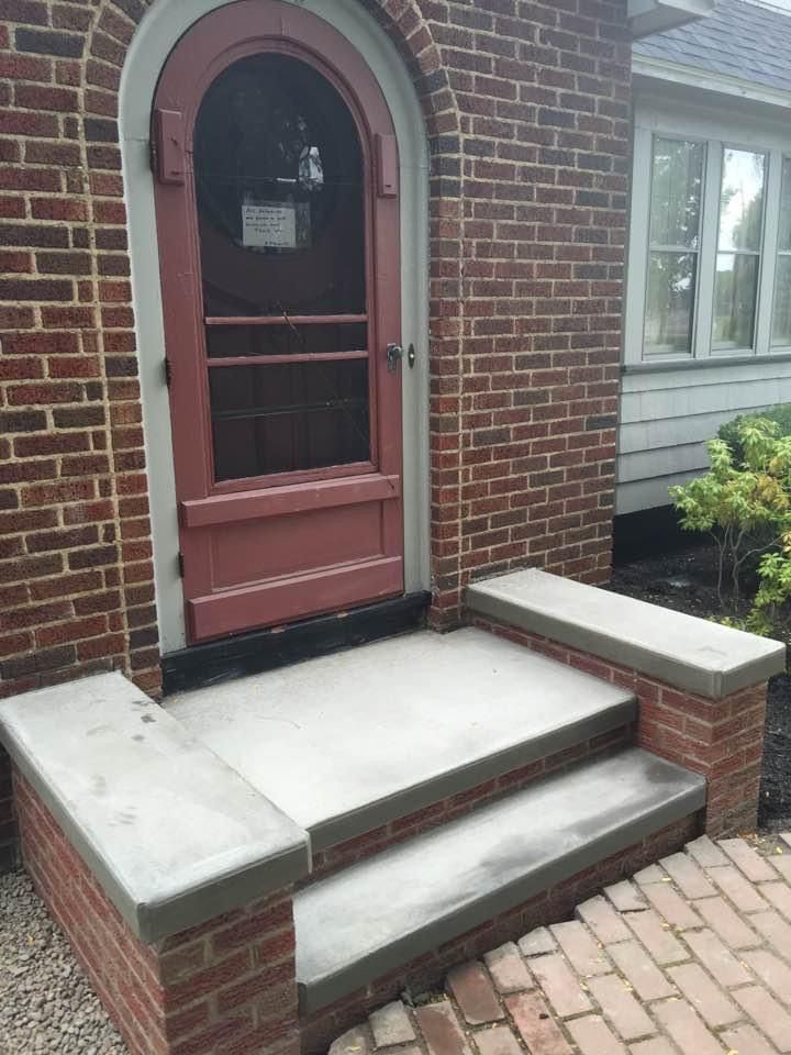 An arched red wooden screen door set in a red brick wall with two concrete steps and matching stone-topped brick side walls.
