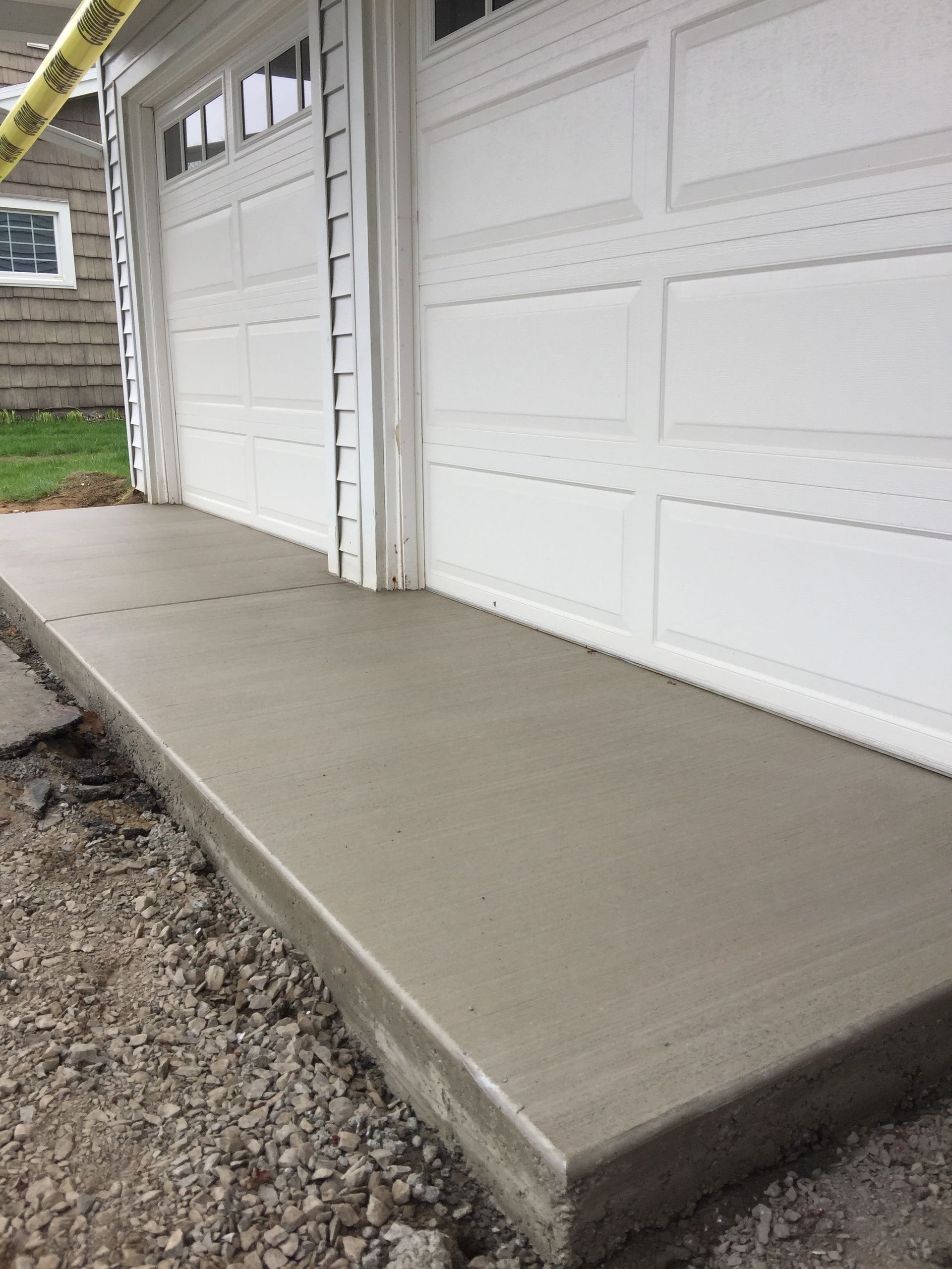 A newly poured concrete slab driveway apron in front of white garage doors, surrounded by gravel and grass.