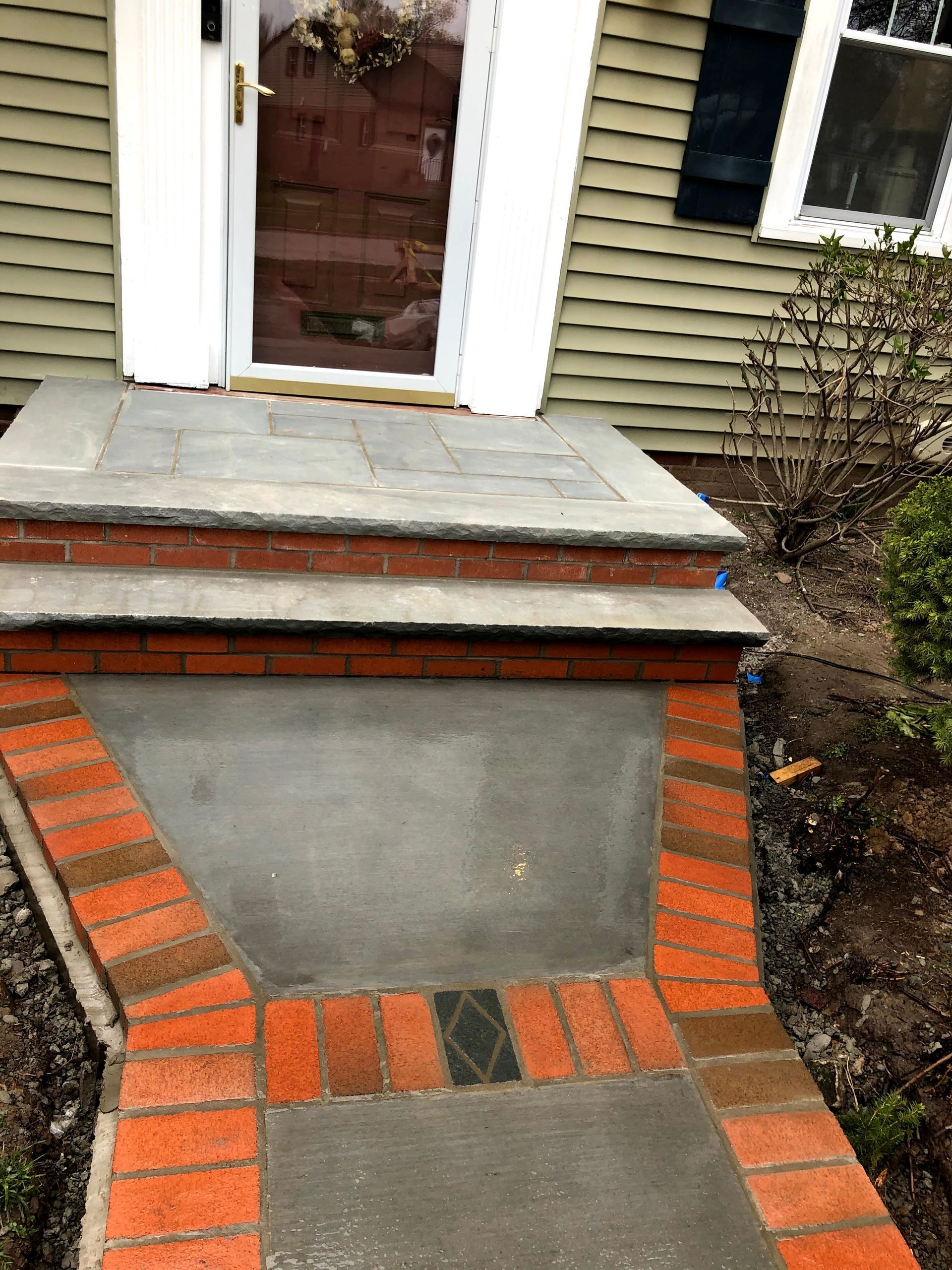A brick and concrete stoop leading to a house entrance, featuring a stone landing, red brick trim, and a concrete path.