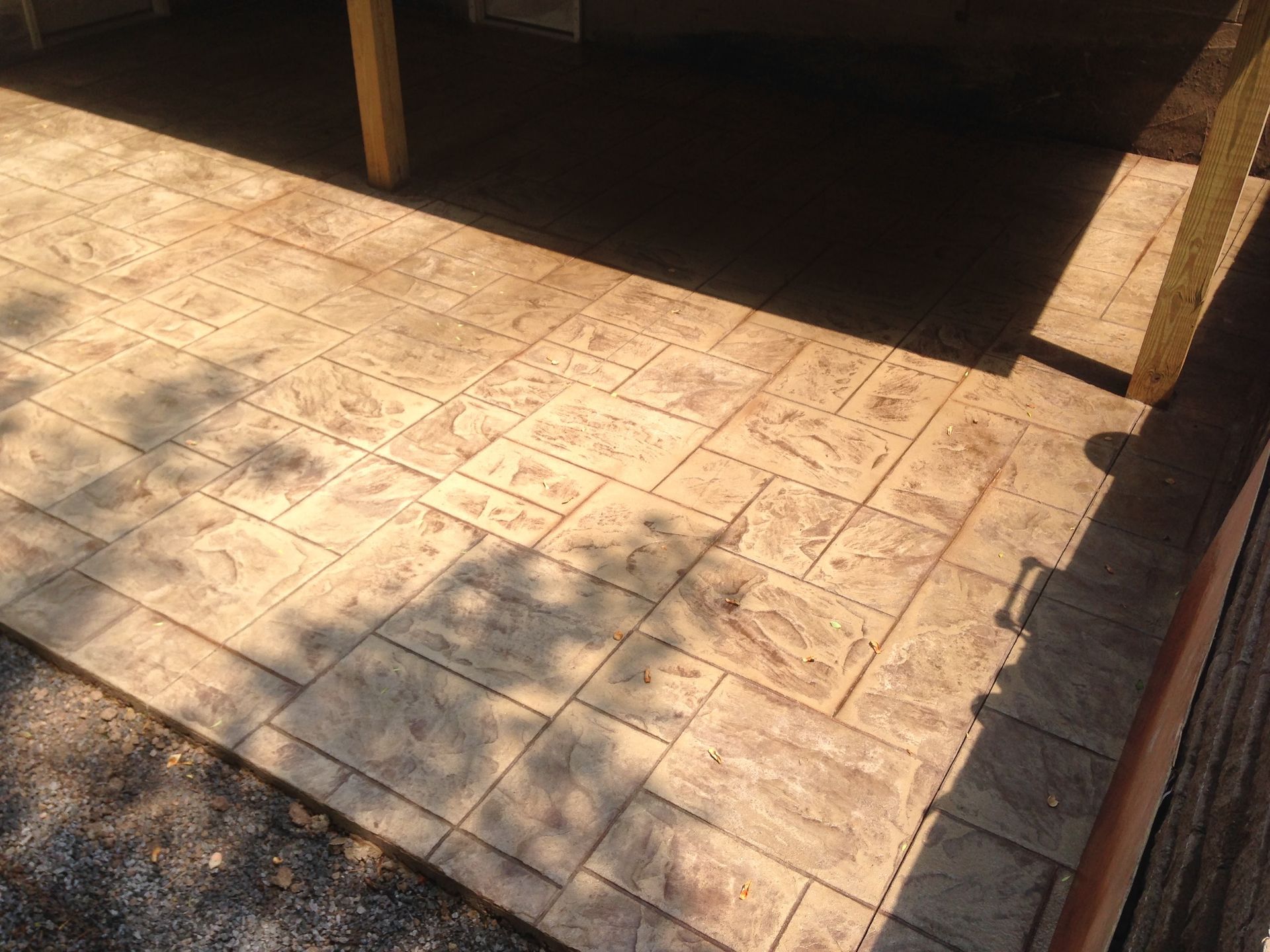 A tan, stamped concrete patio with a rectangular block pattern, partially shaded by a wooden support post.