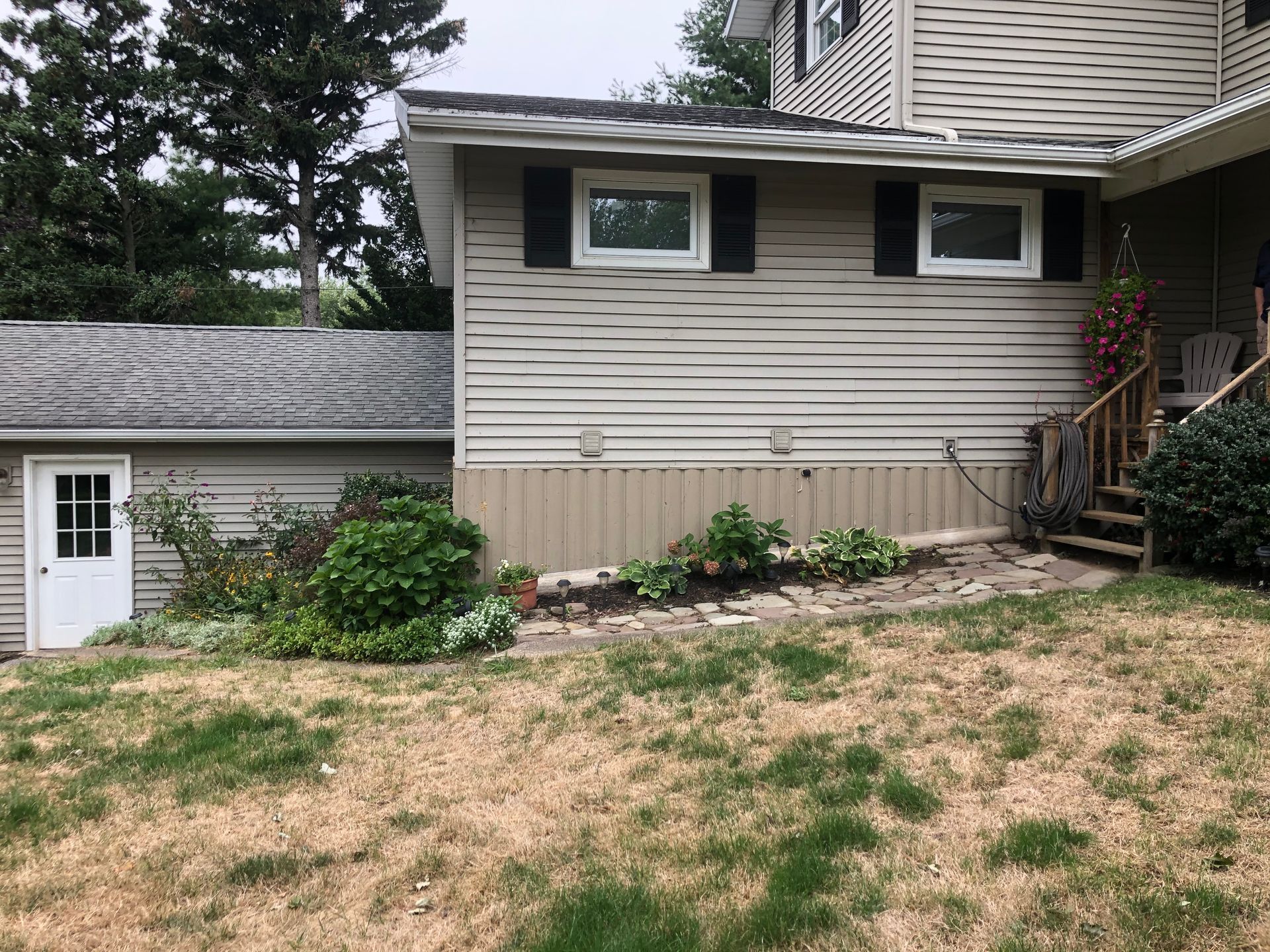 Tan house exterior with vinyl siding, two windows, a white side door, and landscaping against a patchy lawn.
