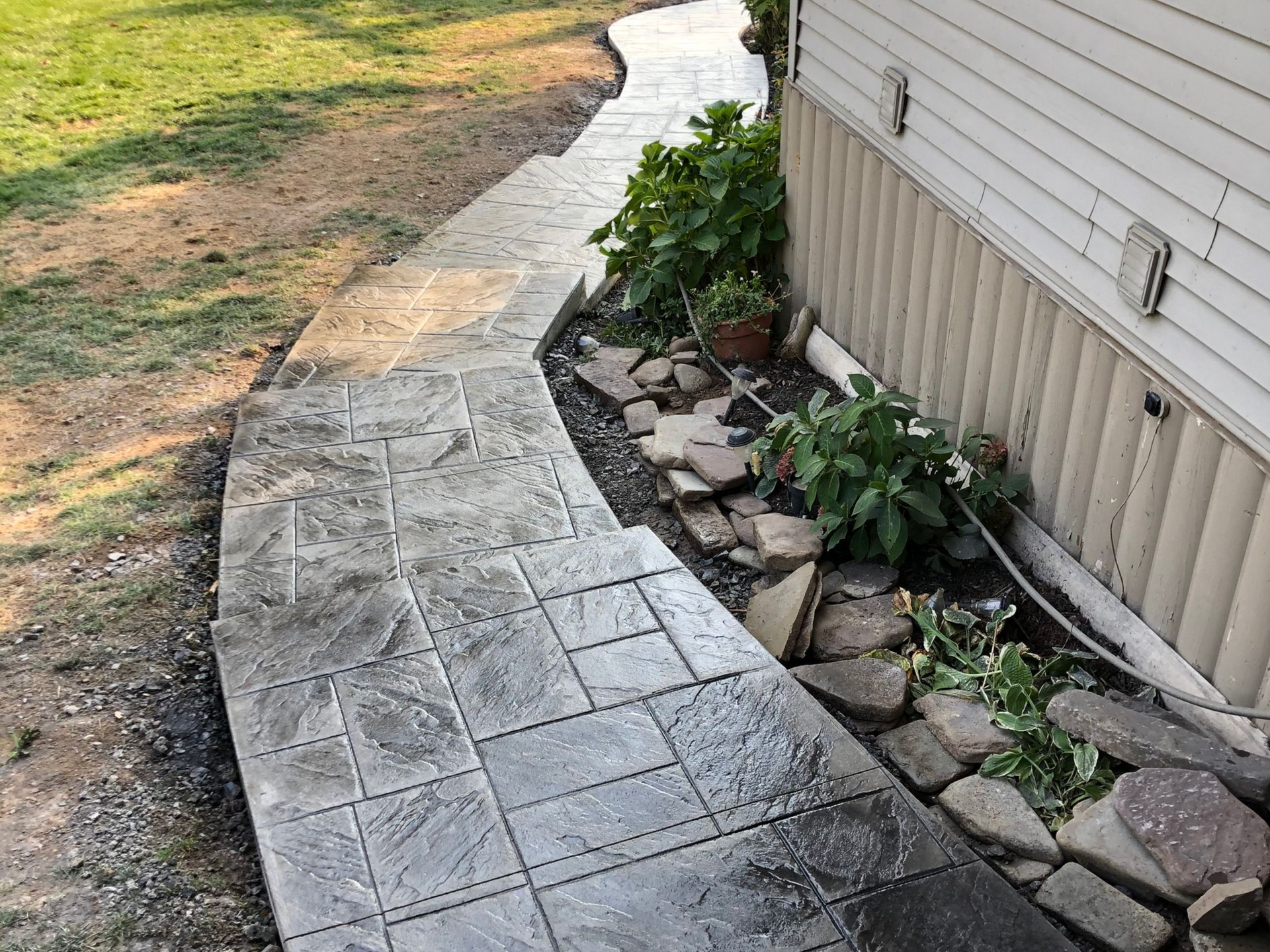 A stamped concrete walkway runs along the side of a building next to a rock garden and a grassy yard.