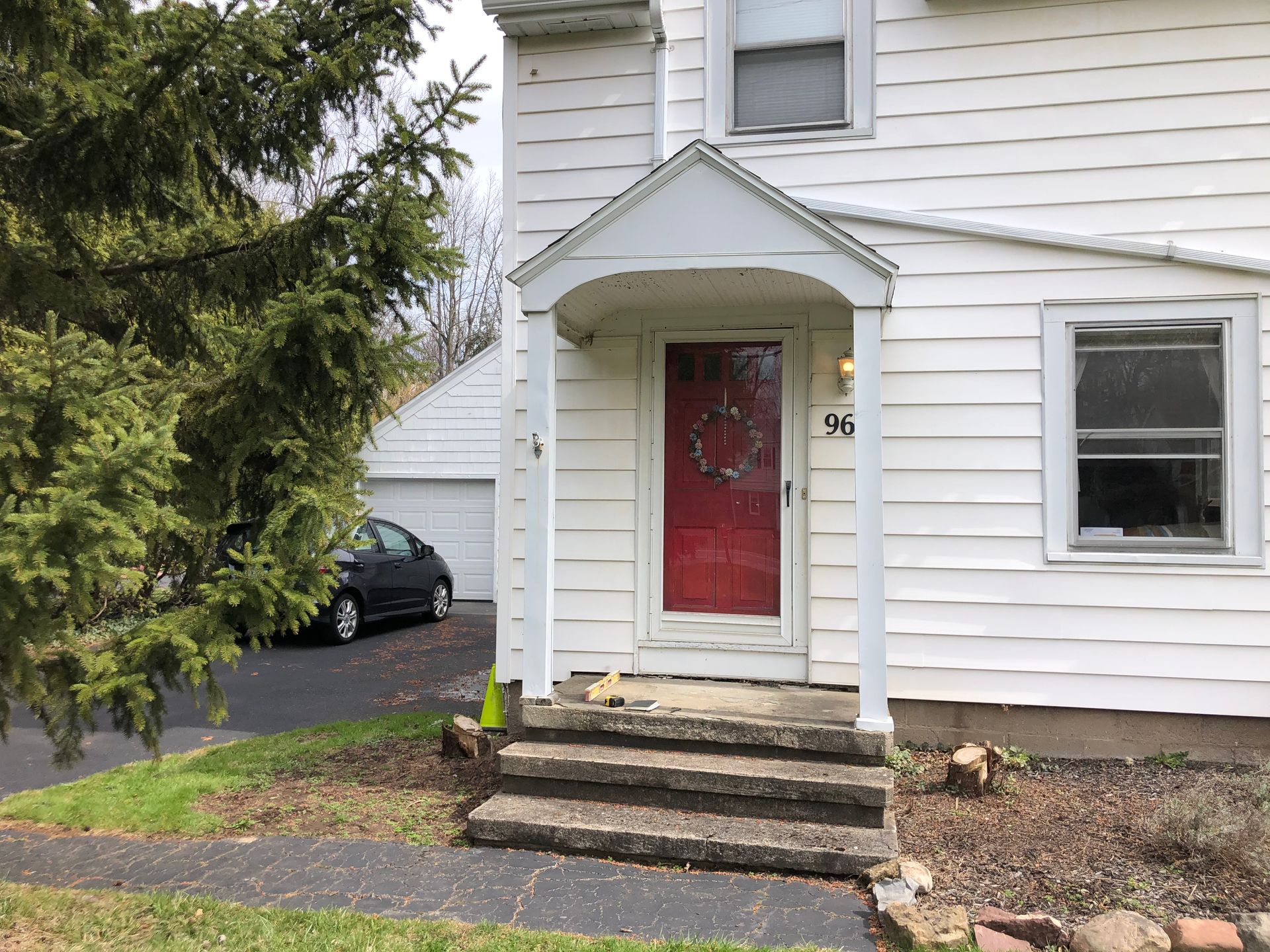 White two-story house with a red front door, concrete steps, and a driveway with a car in the background.