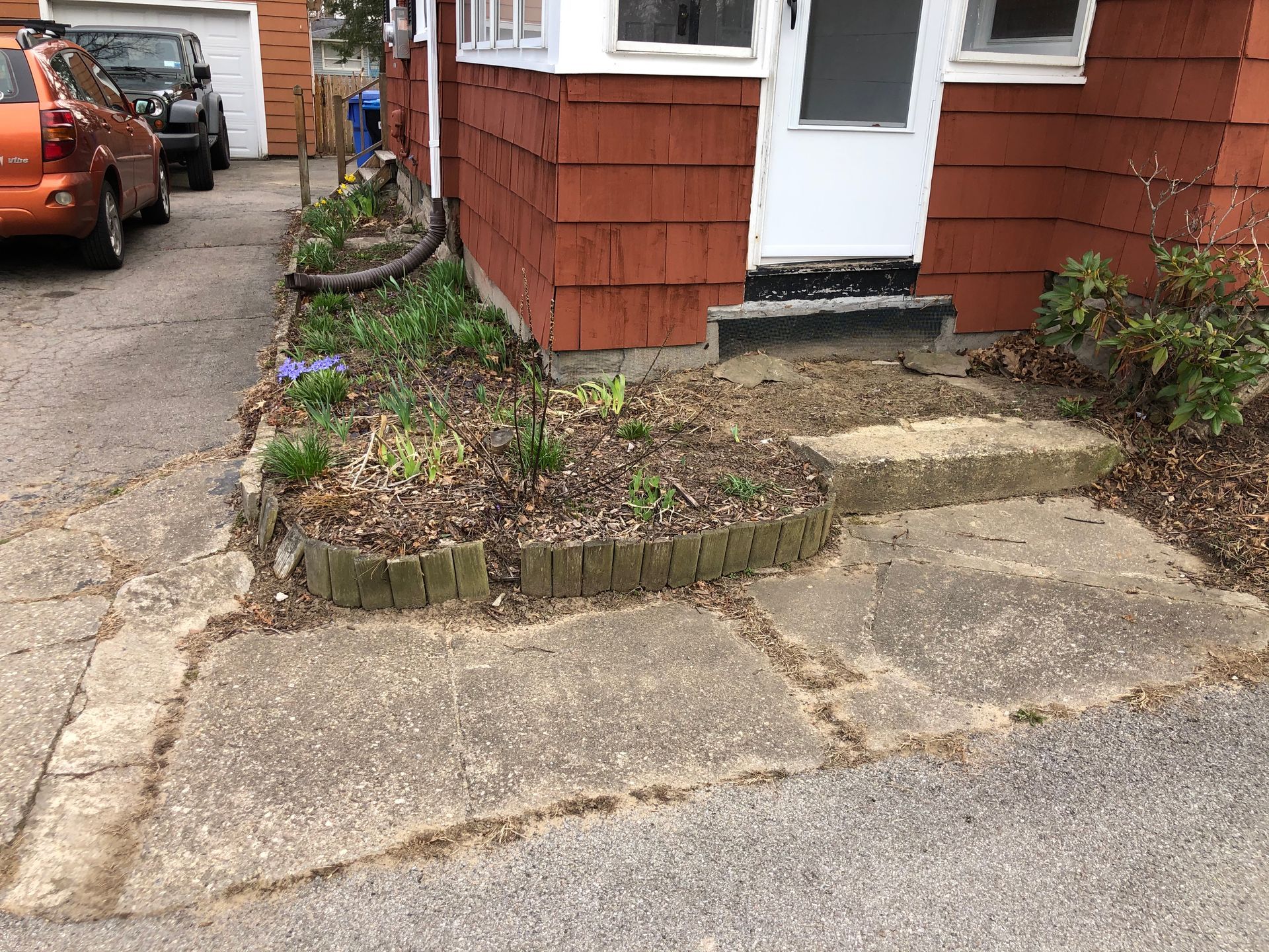A red-shingled house exterior with a small, tiered concrete step leading to a door, next to a raised garden bed and walkway.