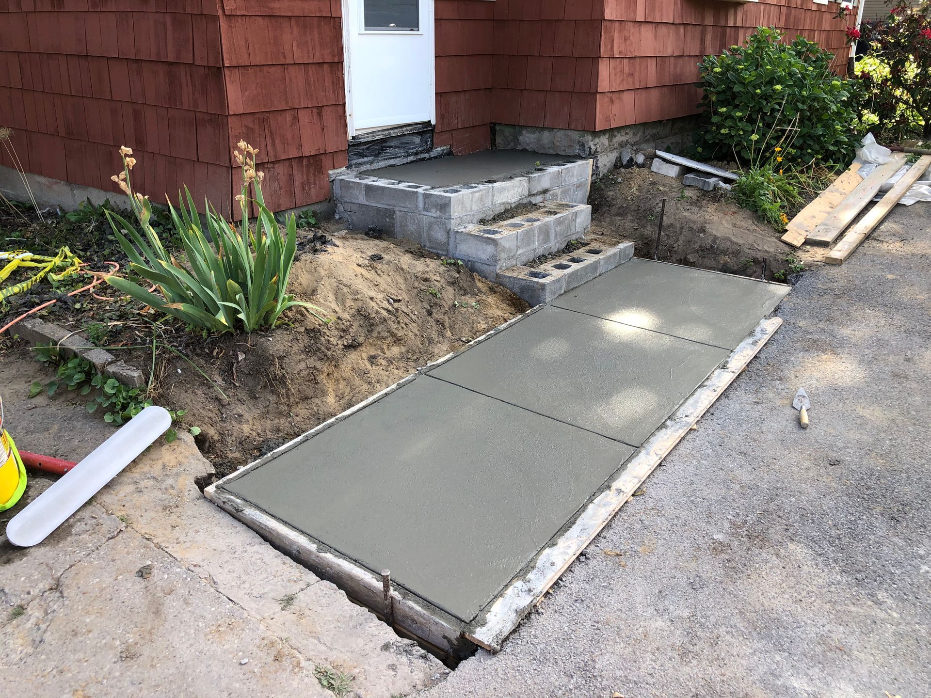 New concrete walkway and step landing construction leading to the front door of a red house with wood shingles.
