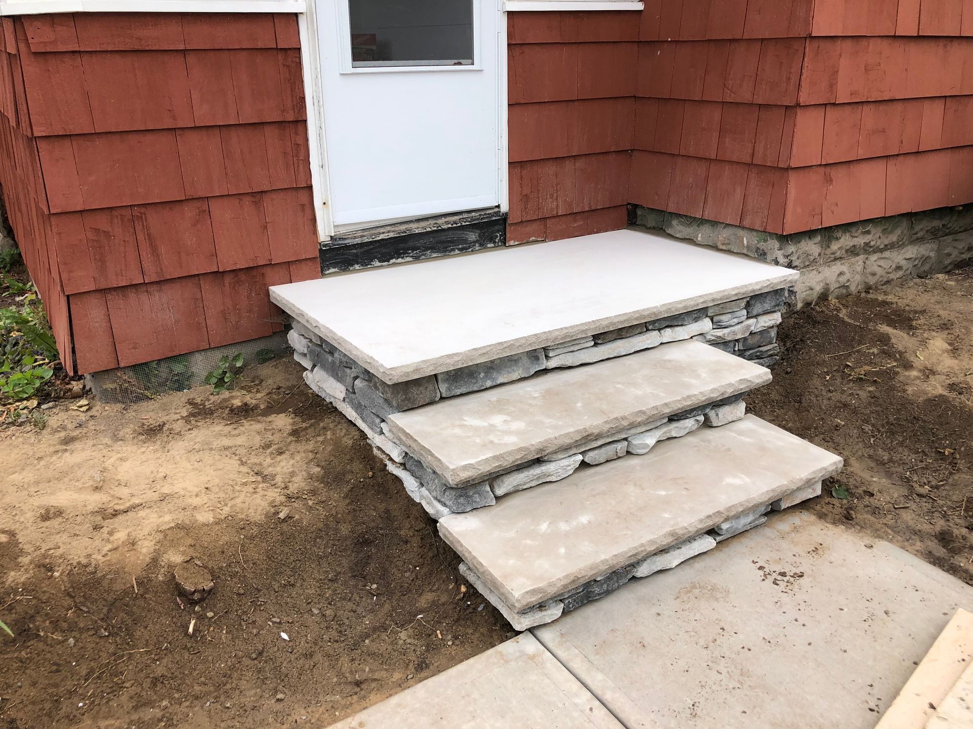 Three stone steps with concrete treads leading to a white exterior door on a house with red siding.
