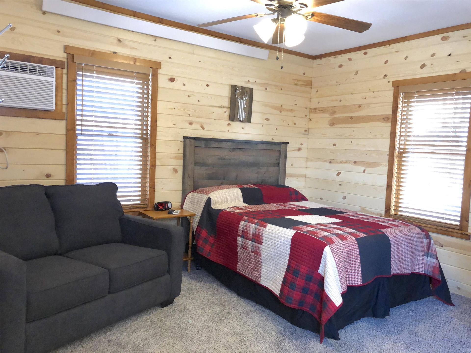 Bedroom with a double bed, gray sofa, wood paneling, and two windows with blinds.