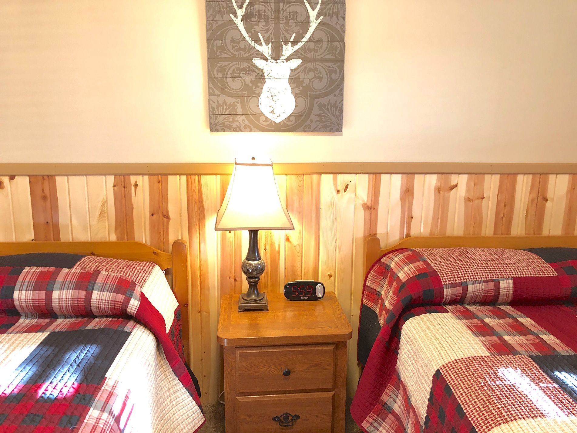 Two twin beds with red plaid quilts and wooden accents, a bedside table with a lamp, and a deer head art piece.