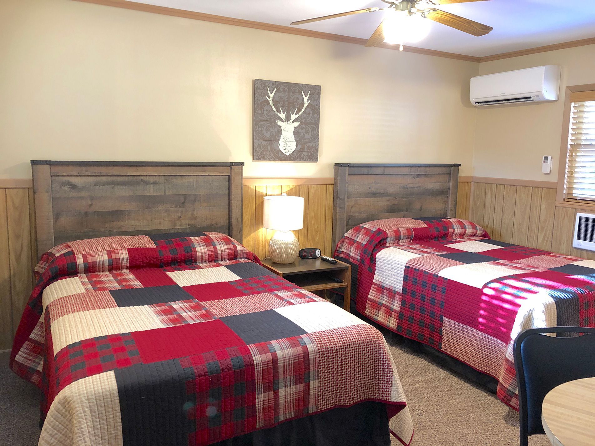Two beds in a room with matching red and gray patchwork quilts, a deer head art, and a small AC unit.