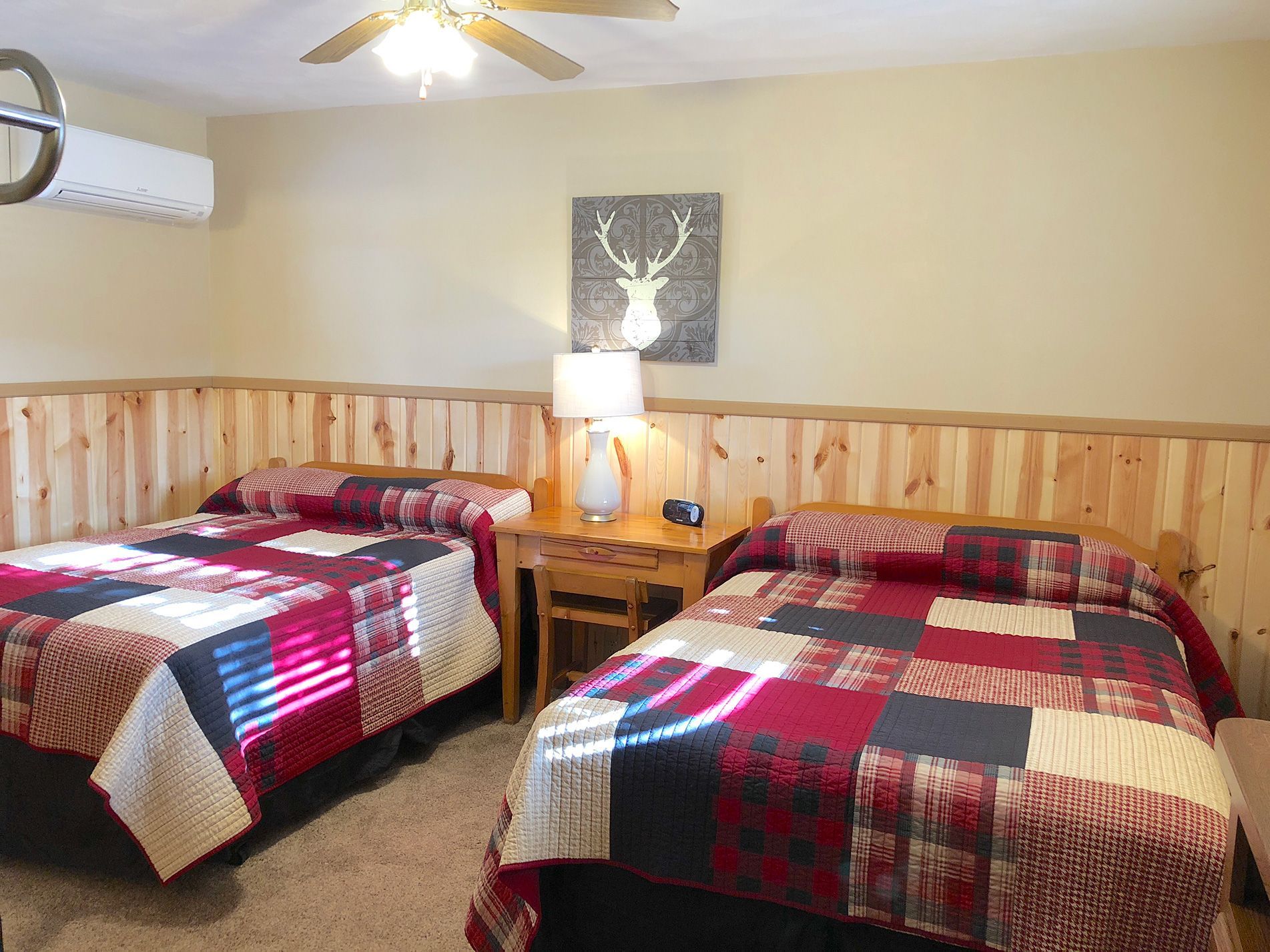 Two beds with red patchwork quilts in a wood-paneled room, a nightstand, and a deer art print.