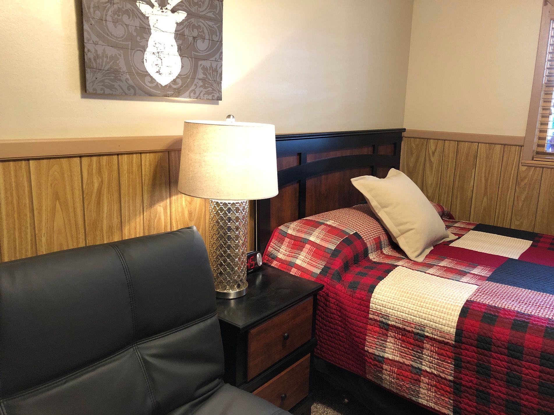 Bedroom with a plaid quilt, lamp, nightstand, and black sofa. Wall art depicts a deer.