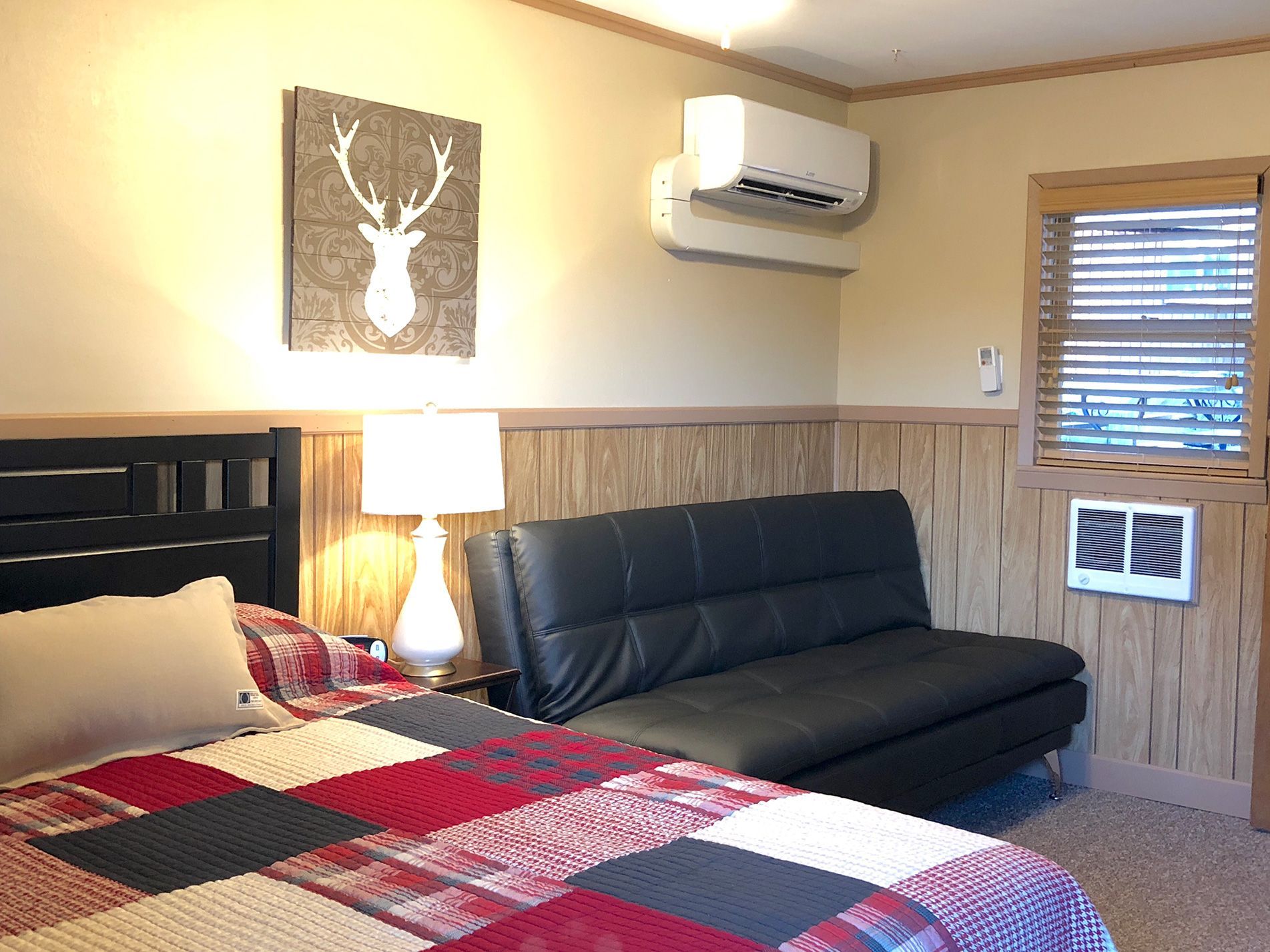 Bedroom with bed, futon, and wall art. Beige walls and paneling, with air conditioner above futon.