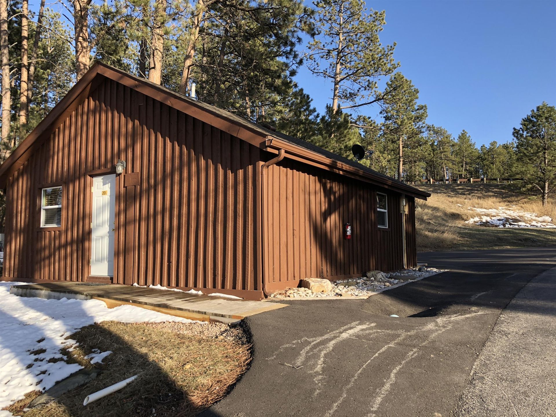 Brown wooden cabin with a paved driveway and patches of snow, set in a wooded area under a blue sky.