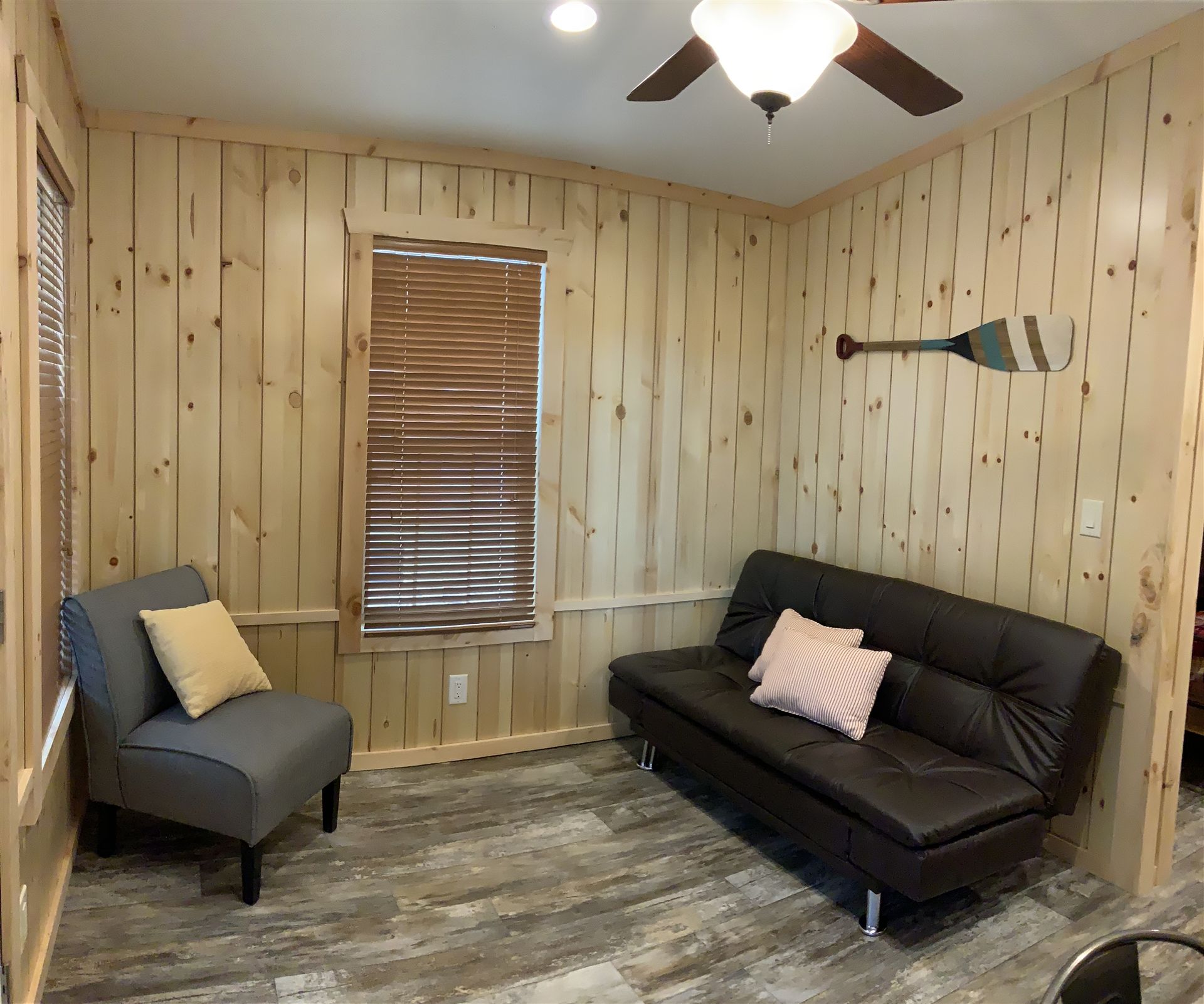 Living room with wood panel walls, gray chair, dark futon, window with blinds, and paddle decor.