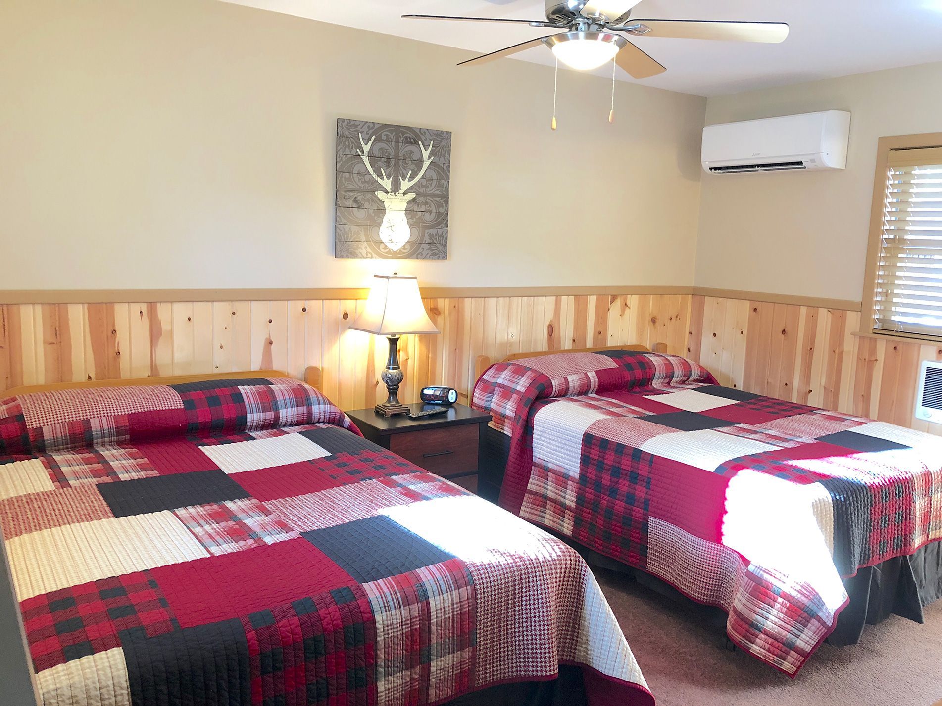 Two beds with red plaid quilts in a room with wood paneling and a deer head art piece.