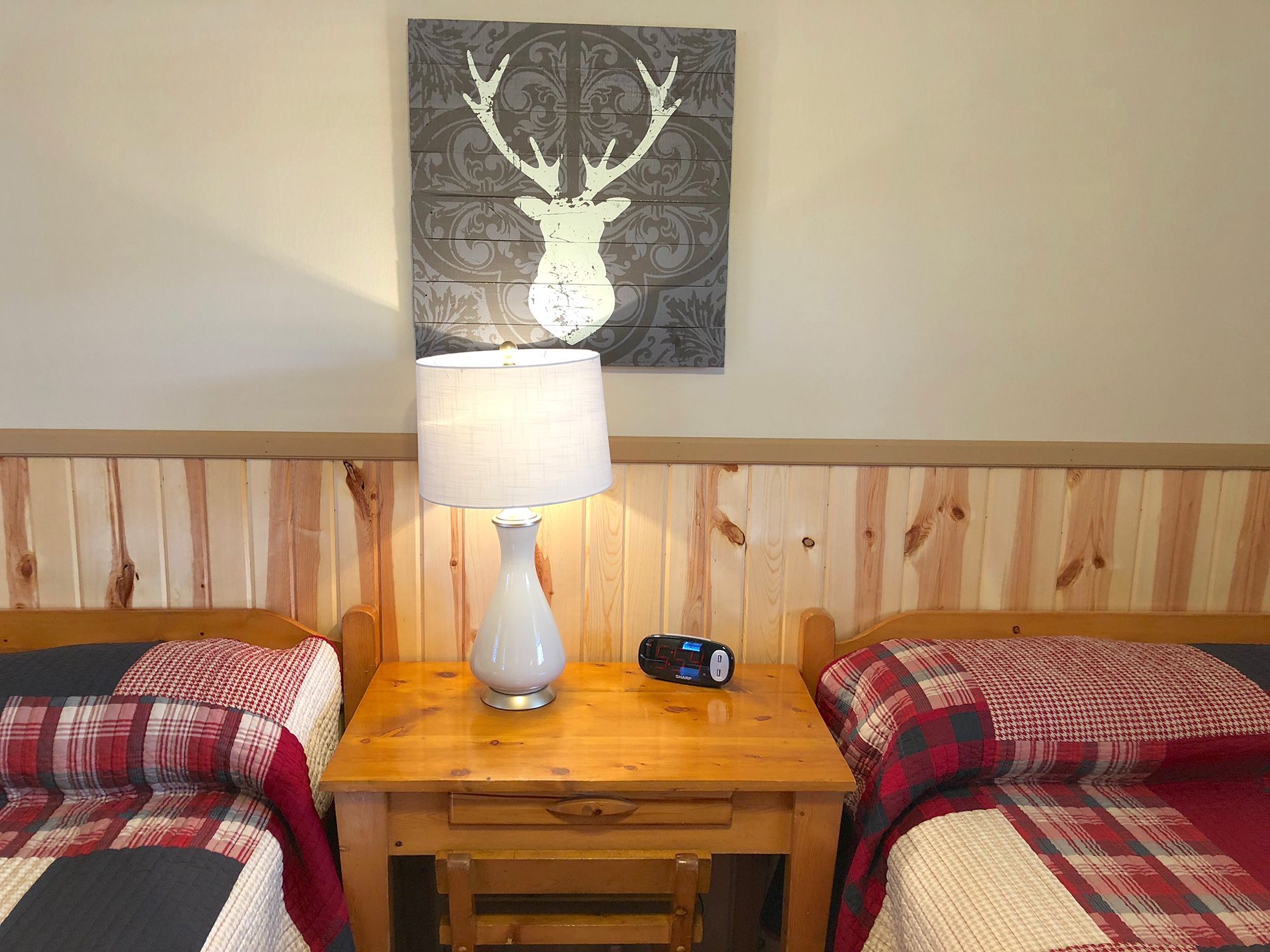 Two twin beds with red plaid bedding flank a wooden nightstand with a lamp. A deer head decor hangs above.