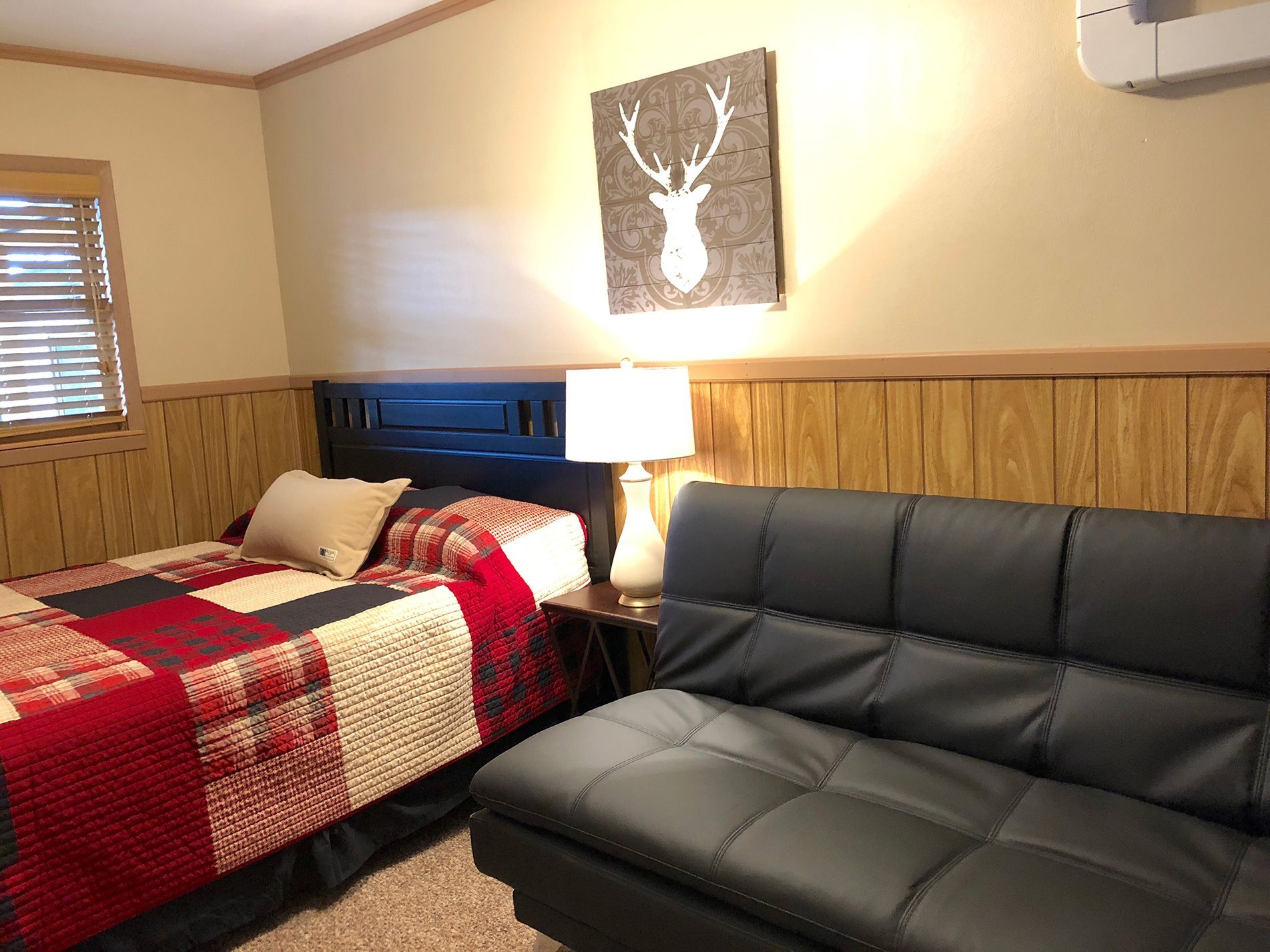 Bedroom with a bed, futon, lamp, and deer art. Red, black, and beige colors. Window with blinds.