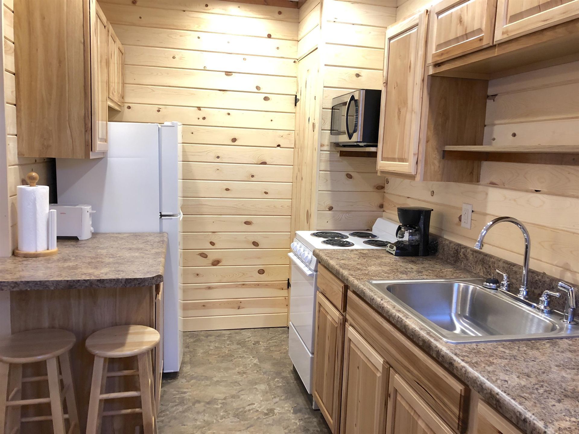 Small kitchen with wooden walls, cabinets, and appliances: refrigerator, stove, microwave, and sink.