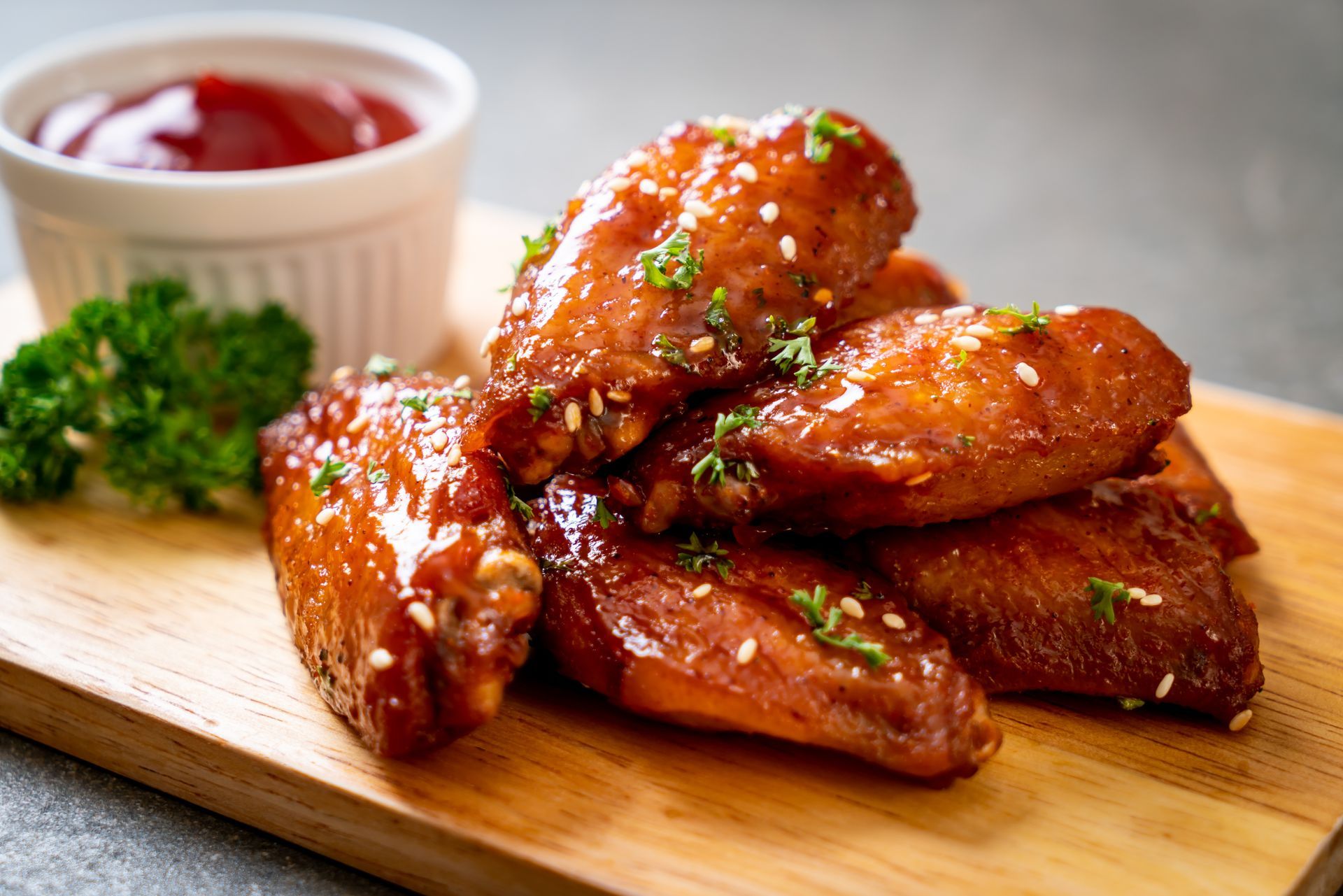 Crispy glazed chicken wings with sesame seeds on a wooden board, served with ketchup and parsley.