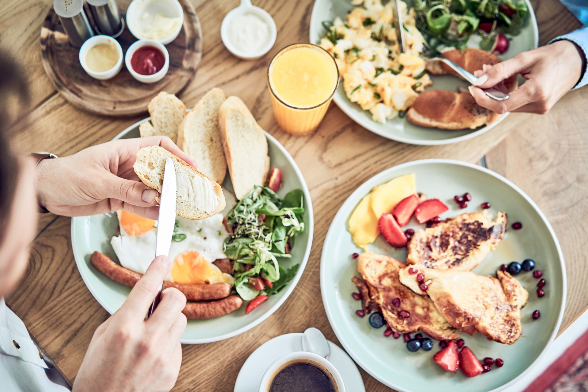 Breakfast table with plates of food, hands buttering toast, and drinks.