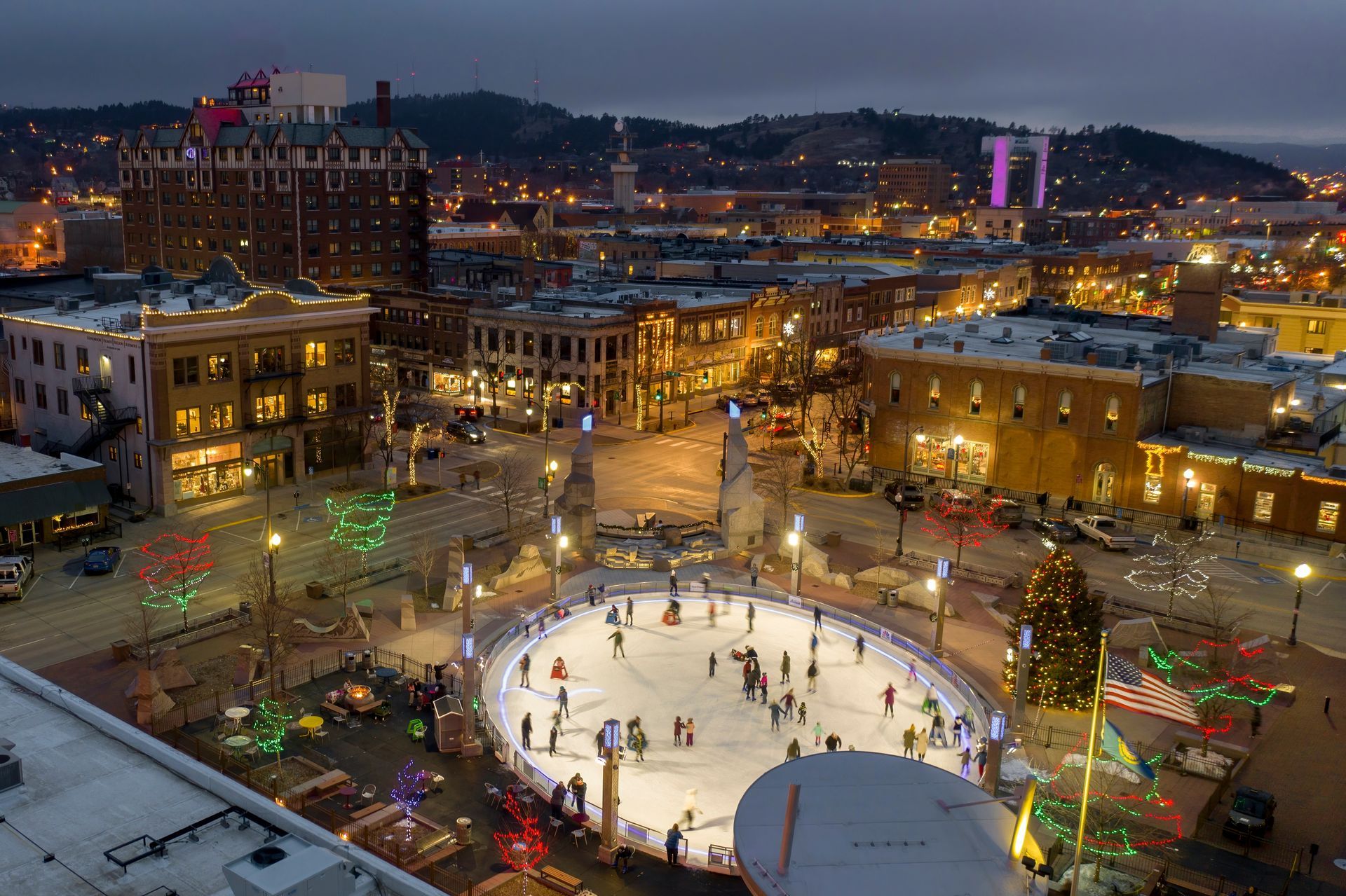 An evening view of a city square with ice skating rink, illuminated buildings, and city lights.