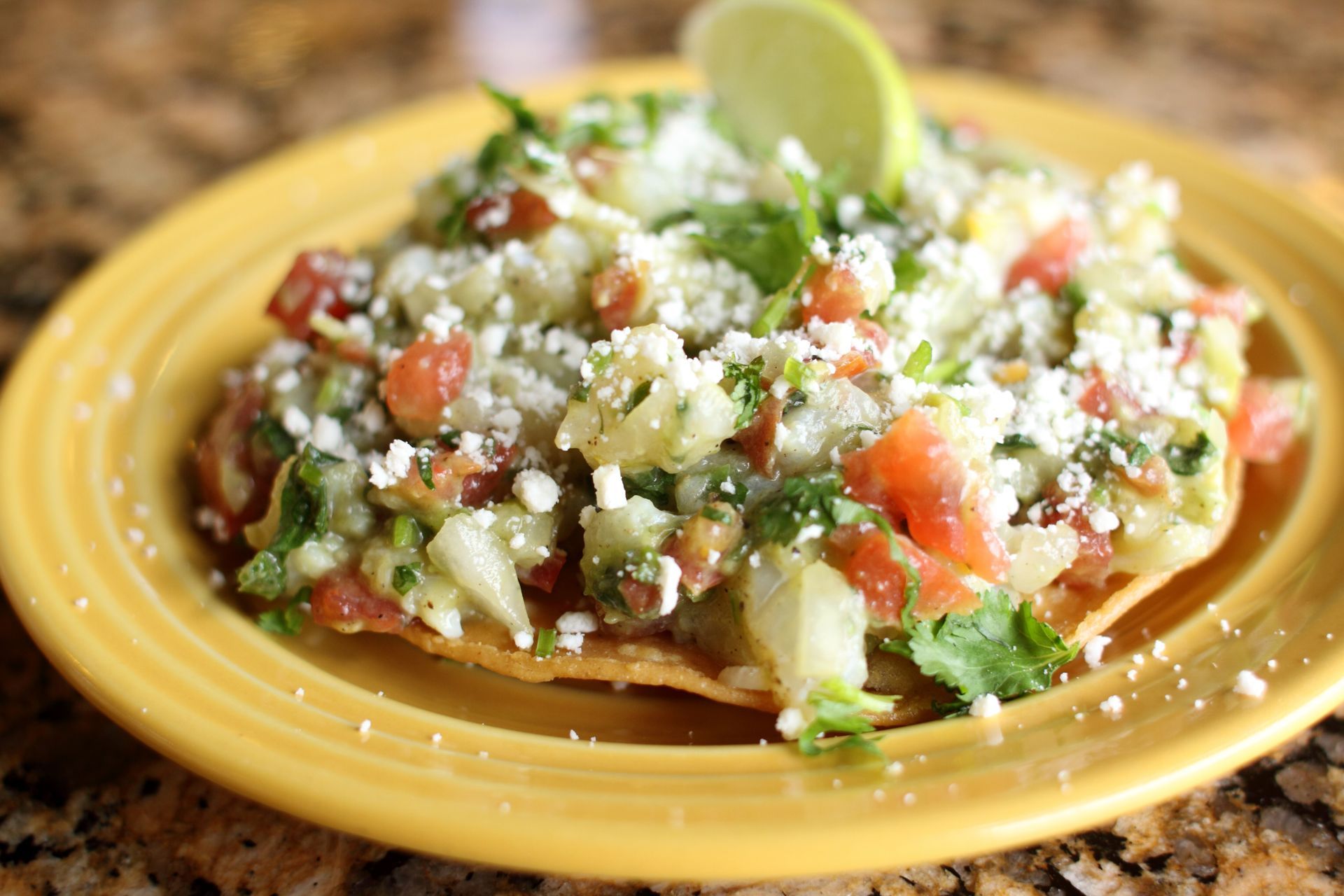 Tostada with chopped tomatoes, cilantro, and cheese, on a yellow plate with lime.