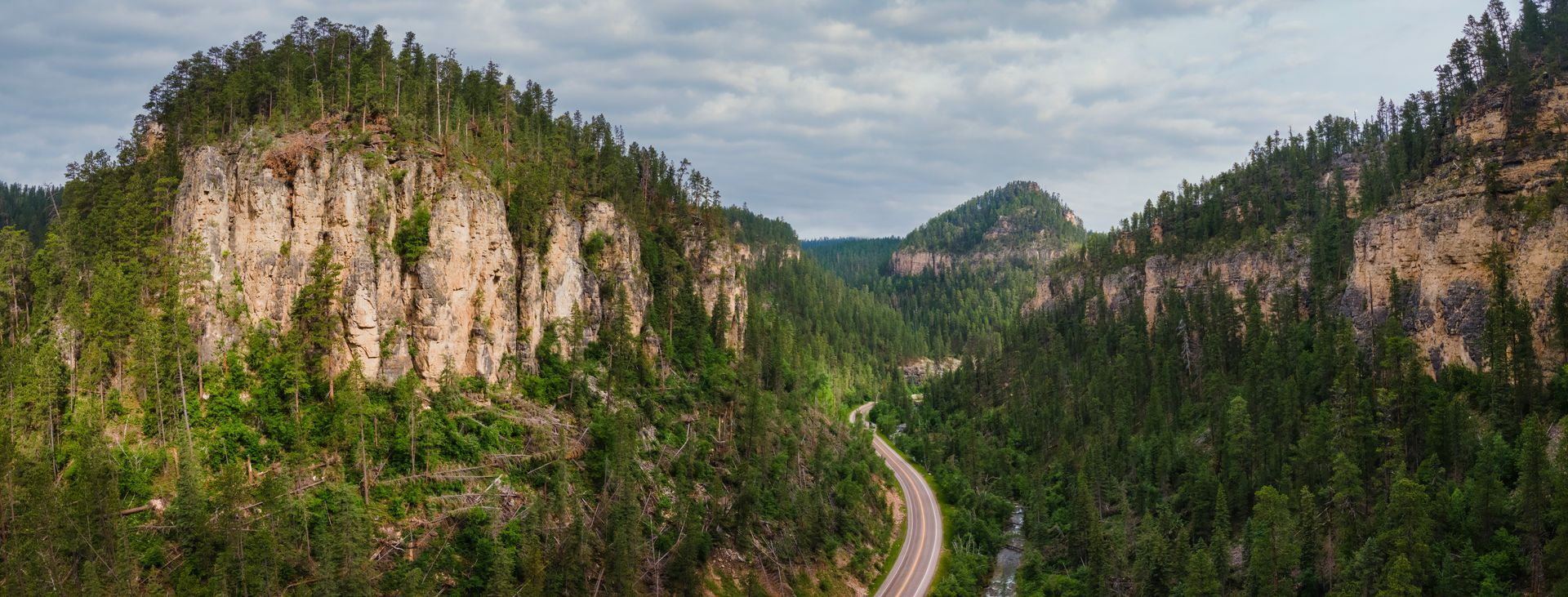 A winding road cuts through a mountainous, forested valley under a cloudy sky.