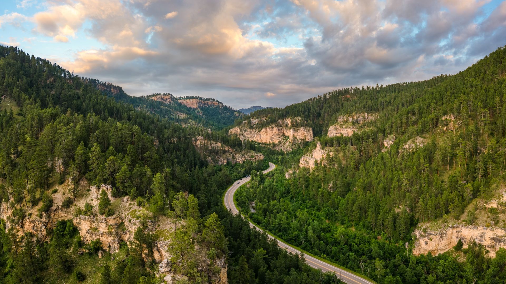Winding road through a green forested mountain valley under a cloudy sky.