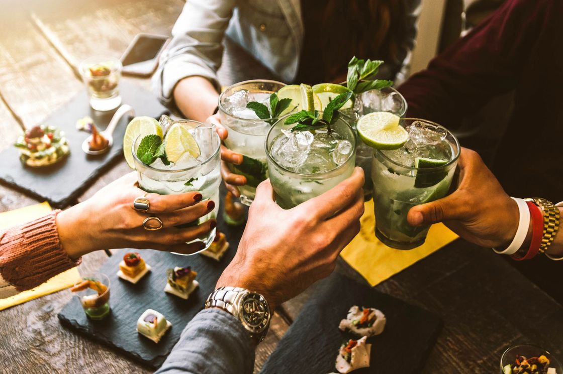 Friends toasting with cocktails; lime wedges, mint, and ice in glasses, appetizers on slate boards.