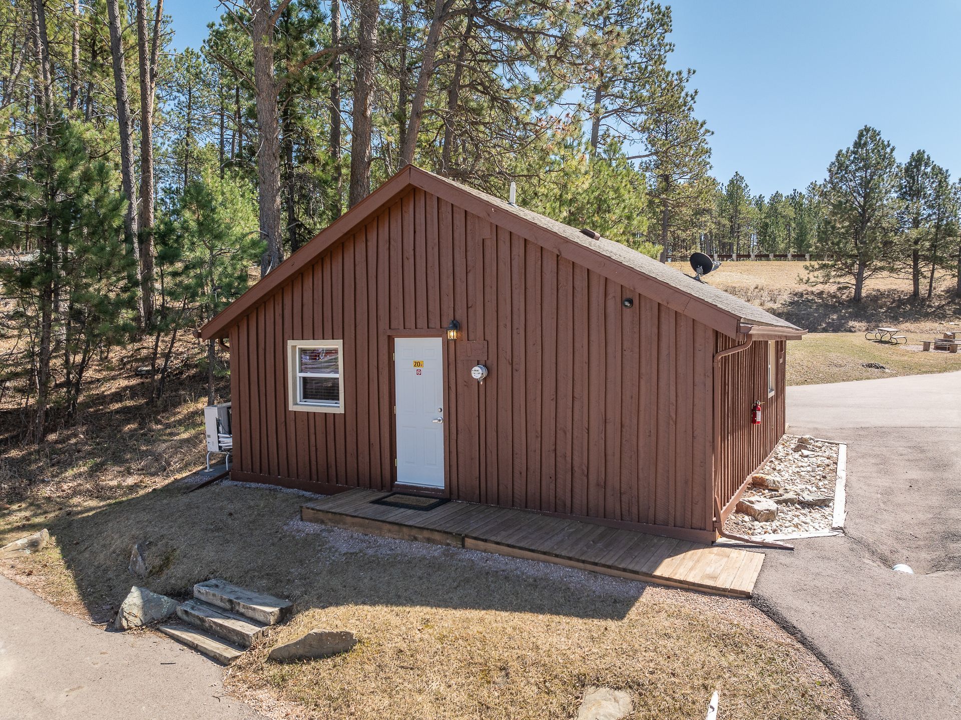 Brown cabin with white door and window, set in a wooded area with a paved driveway.