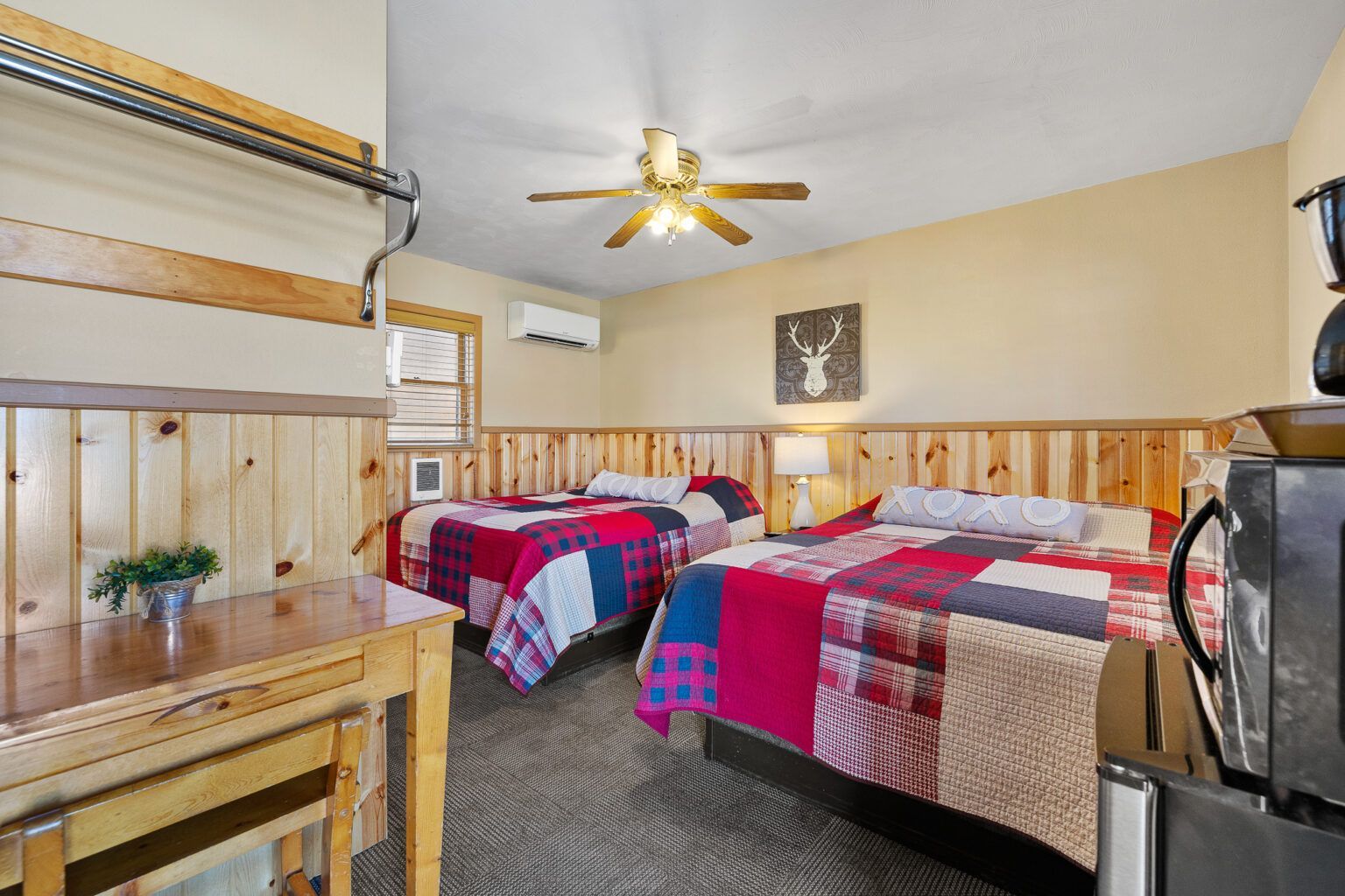 Bedroom with two beds, wood paneling, and a desk, featuring colorful quilts and a ceiling fan.