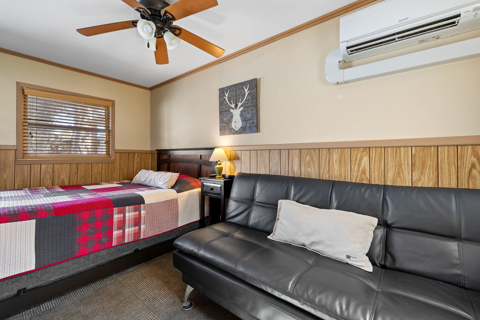 Small bedroom with bed, black futon, wall art, and air conditioner. Brown wood paneling.