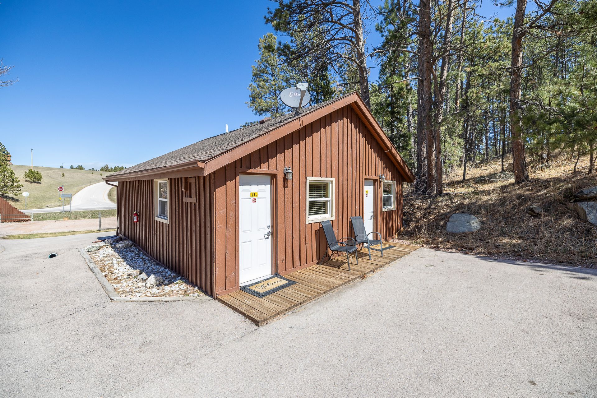 Brown cabin with white doors and windows, gravel driveway, surrounded by trees under a blue sky.