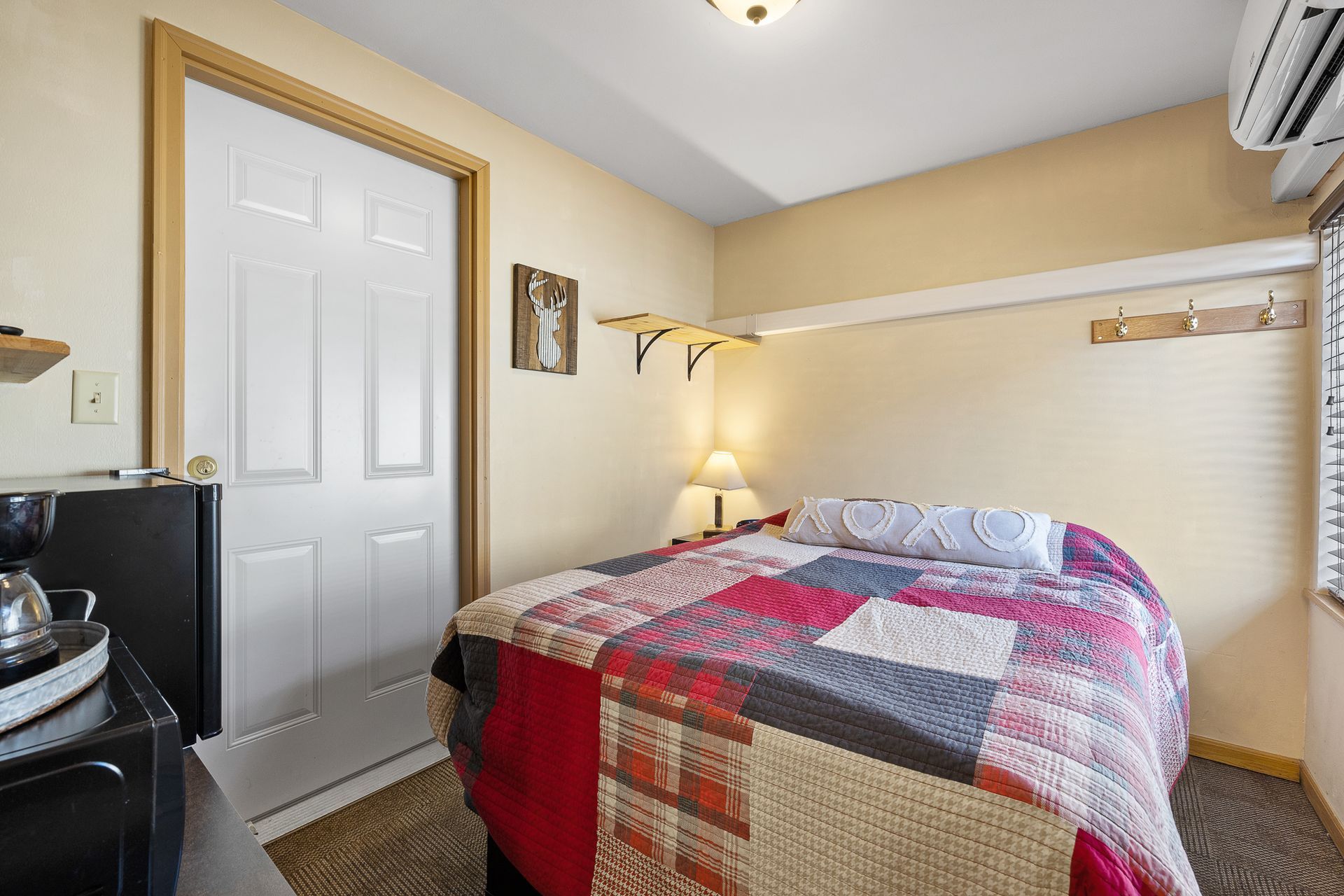 Small bedroom with bed, door, and mini-fridge. Patchwork quilt, beige walls, and window with blinds.