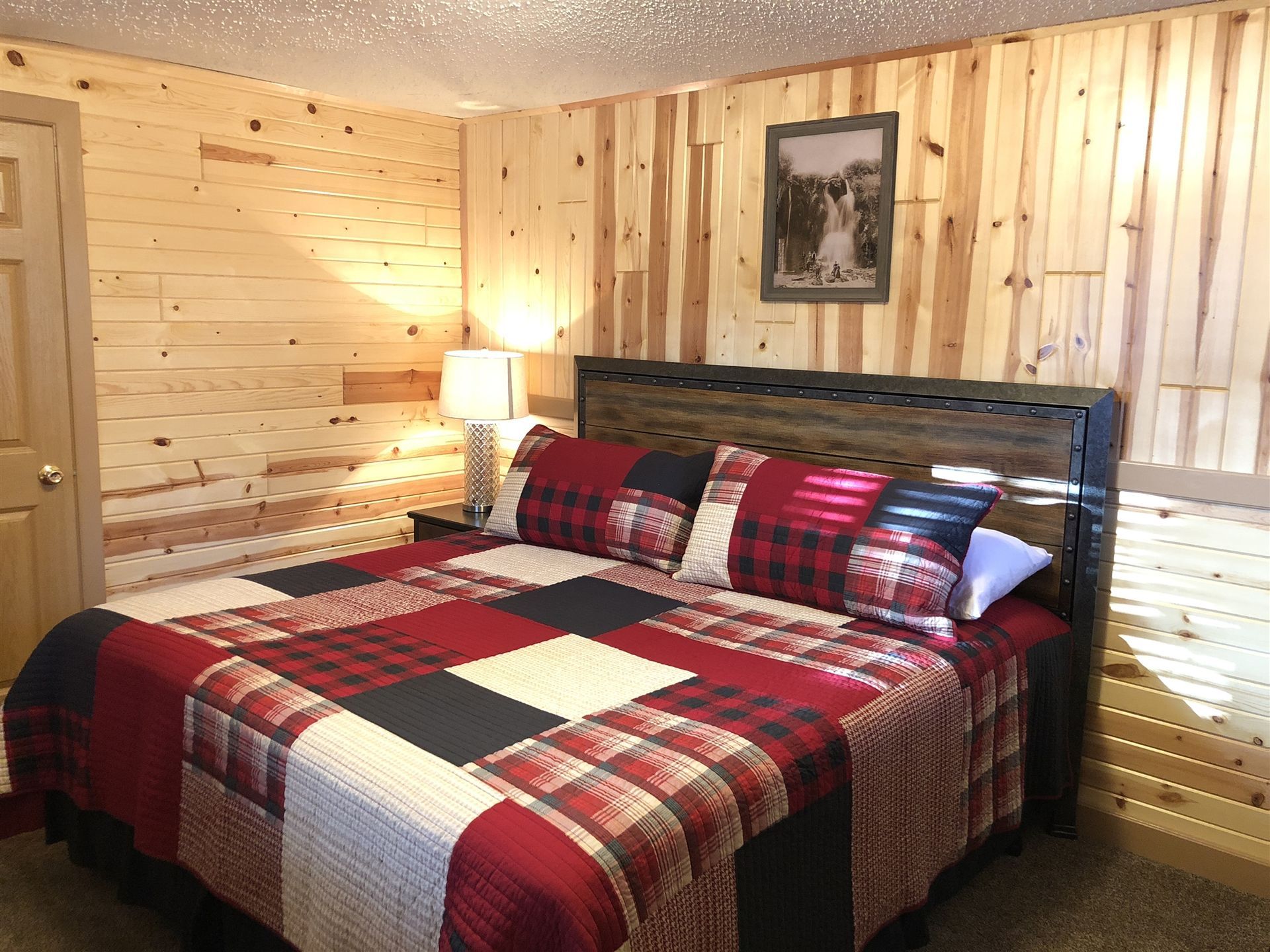 Bedroom with wood-paneled walls, a king-sized bed with a plaid quilt, and a framed picture.