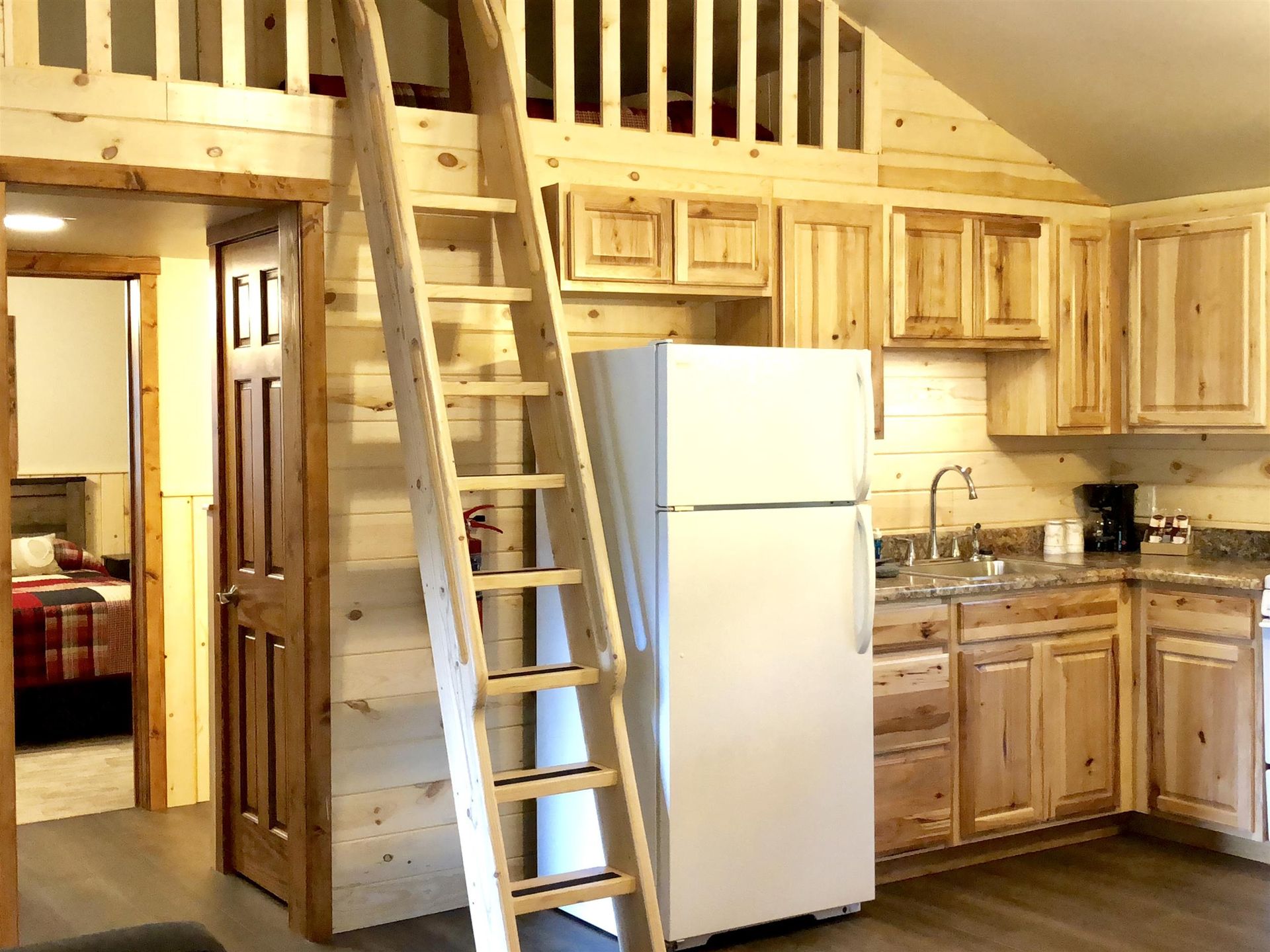 Cabin interior with loft, ladder, kitchen, and open doorway to bedroom. Wooden walls and cabinets.