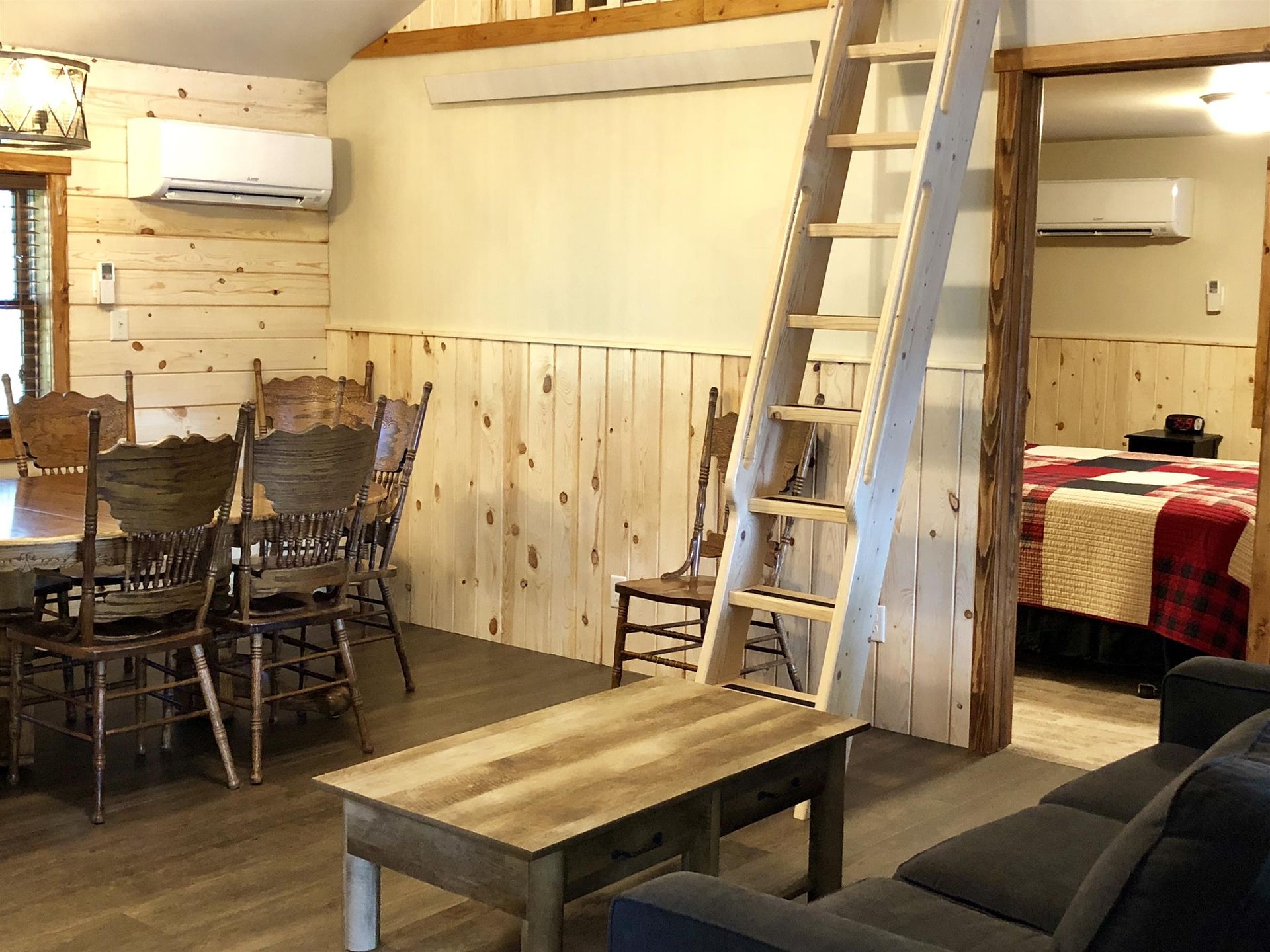 Interior cabin with dining area, living space with a couch, and bedroom visible; a ladder leads up.