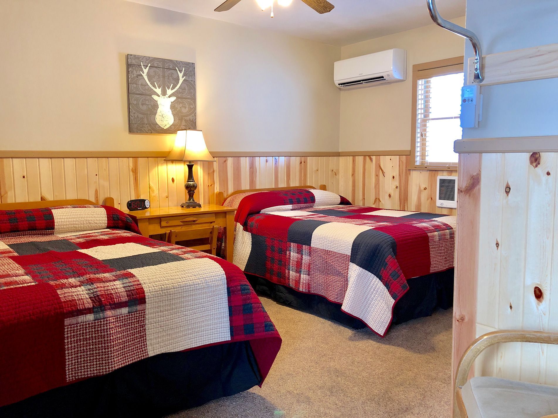 Two-bed motel room with red and black quilts, a wooden accent wall, and a deer head art piece.